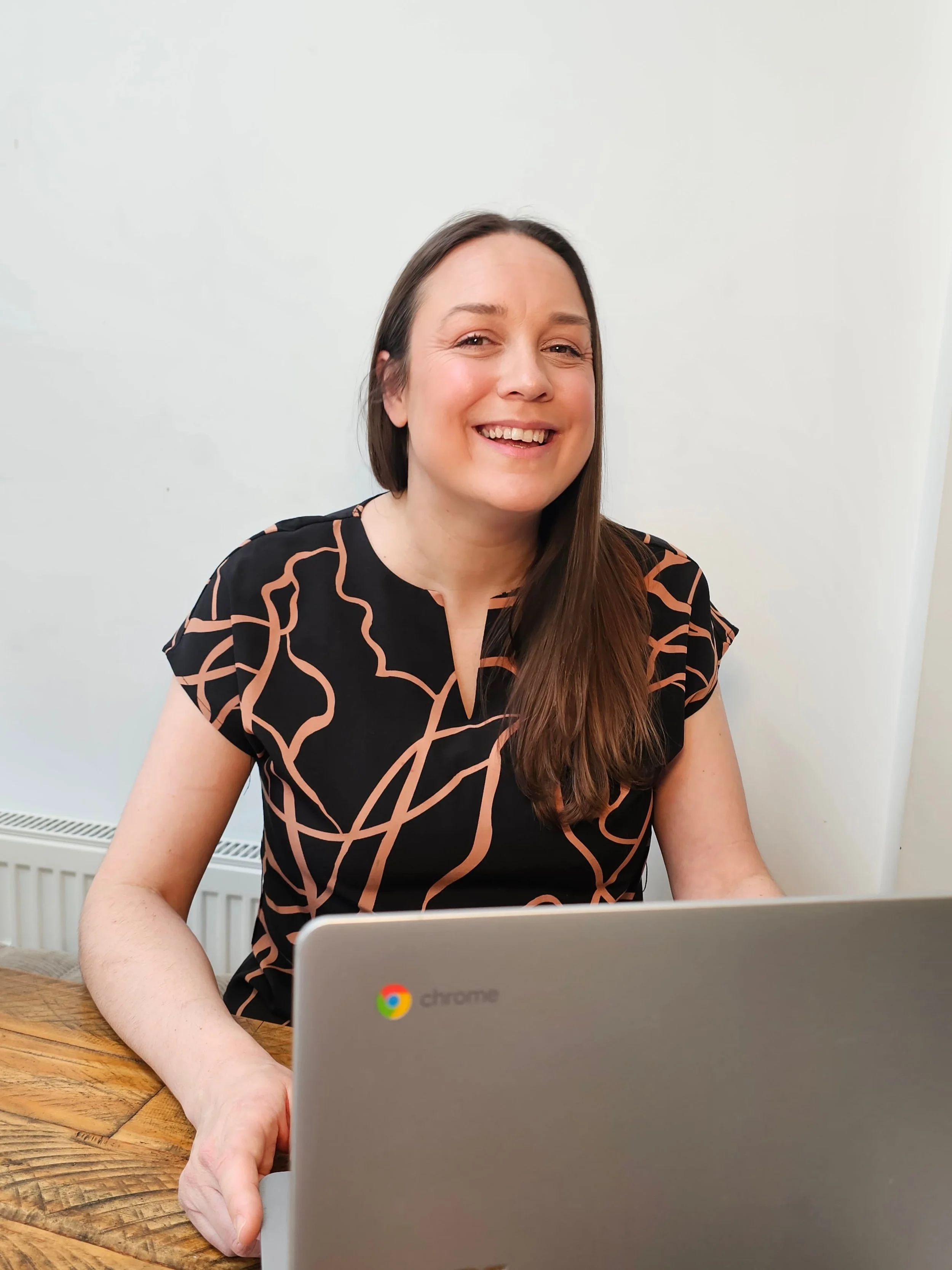 A smiling woman with long brown hair working on a laptop at a wooden table against a plain white wall.