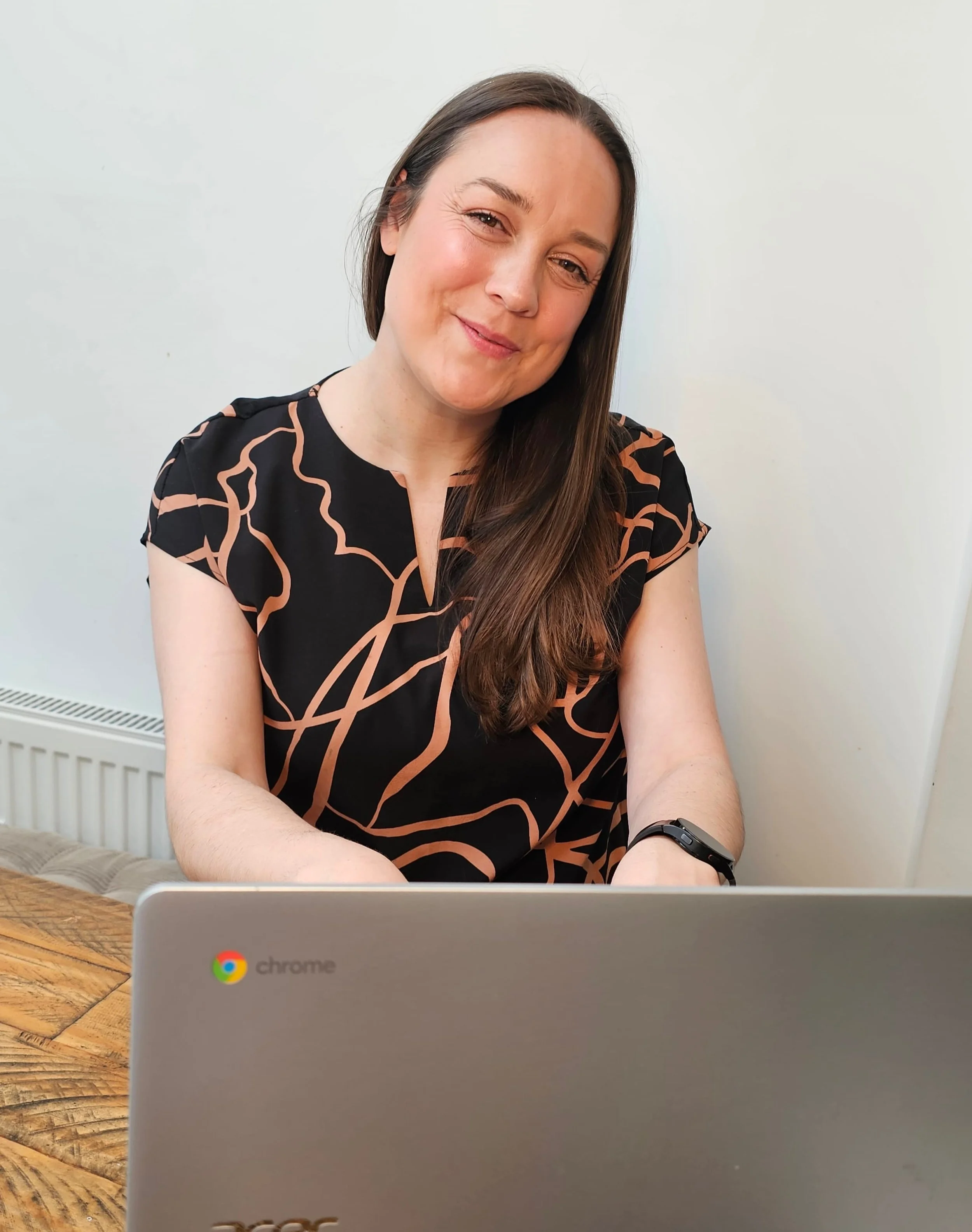 A woman with long brown hair, smiling, sitting at a table with a laptop in front of her, wearing a black dress with a tan abstract pattern, in a room with white walls.