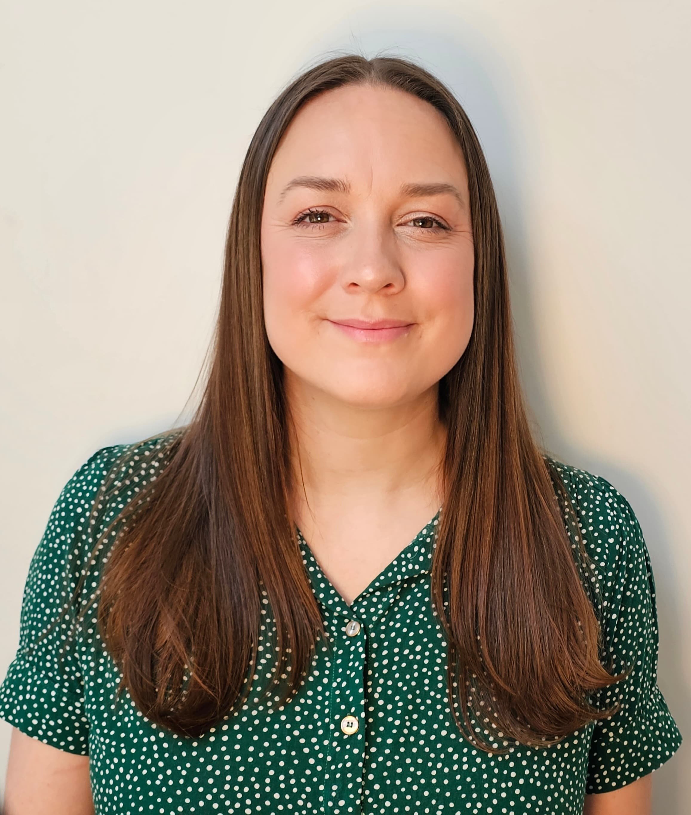 A woman with long brown hair smiling at the camera, wearing a green blouse with white polka dots, standing against a plain light-colored background.