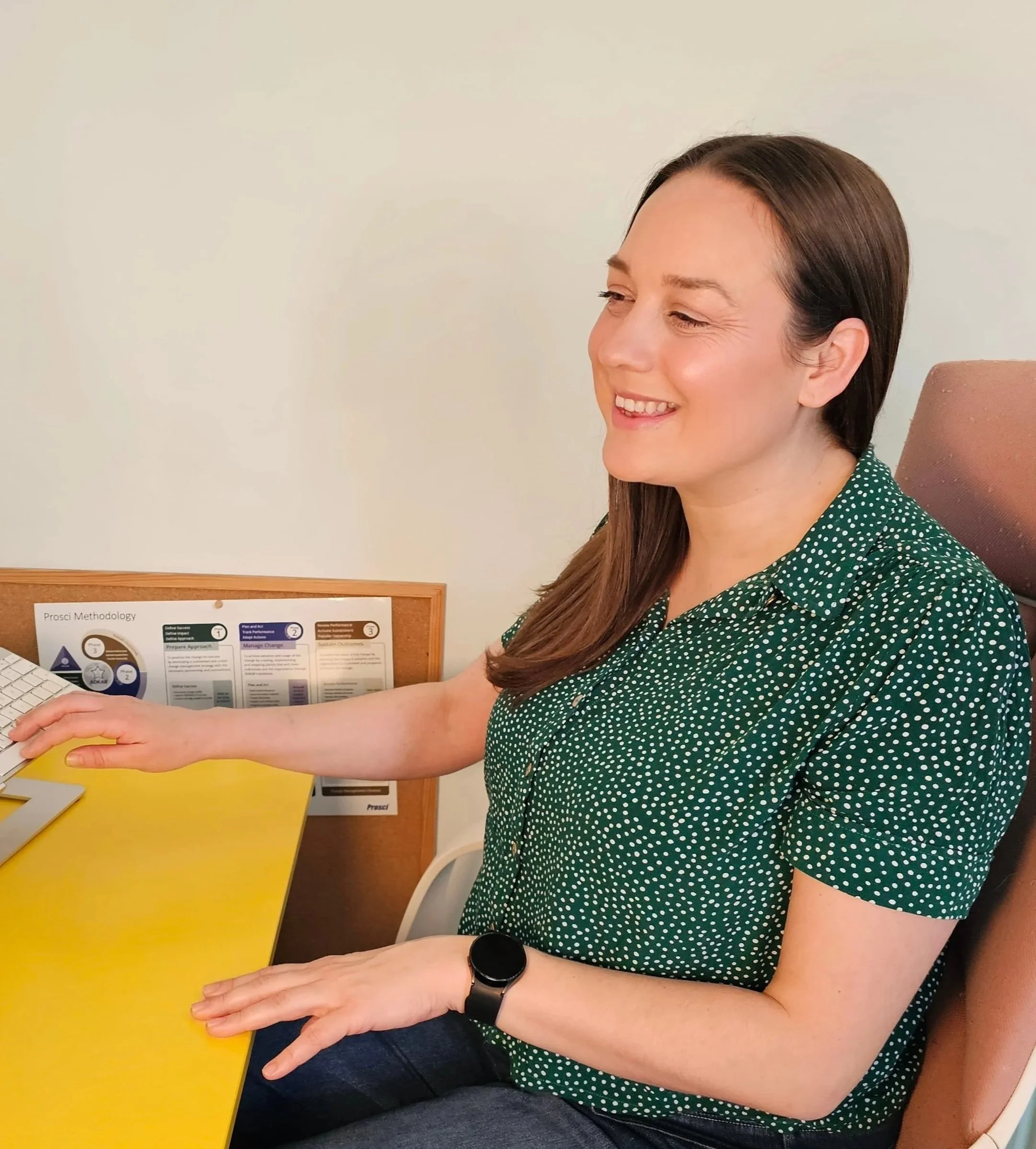 A woman sitting at a yellow desk, wearing a green polka dot shirt, smiling while working on a computer in an office setting.