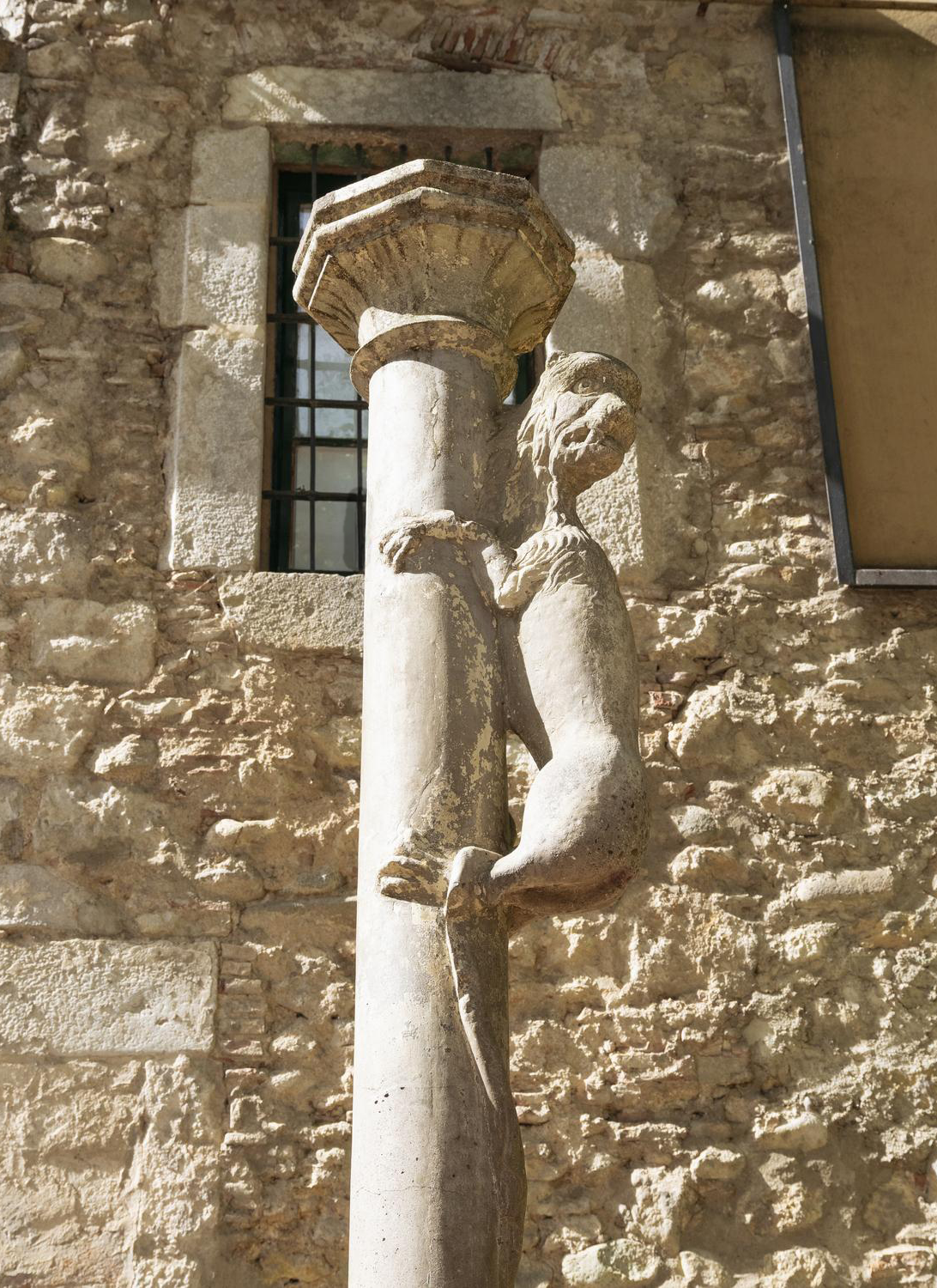Sculpture of a humanoid figure with a bird-like head clinging to a stone column, set against a background of a stone wall with a window.