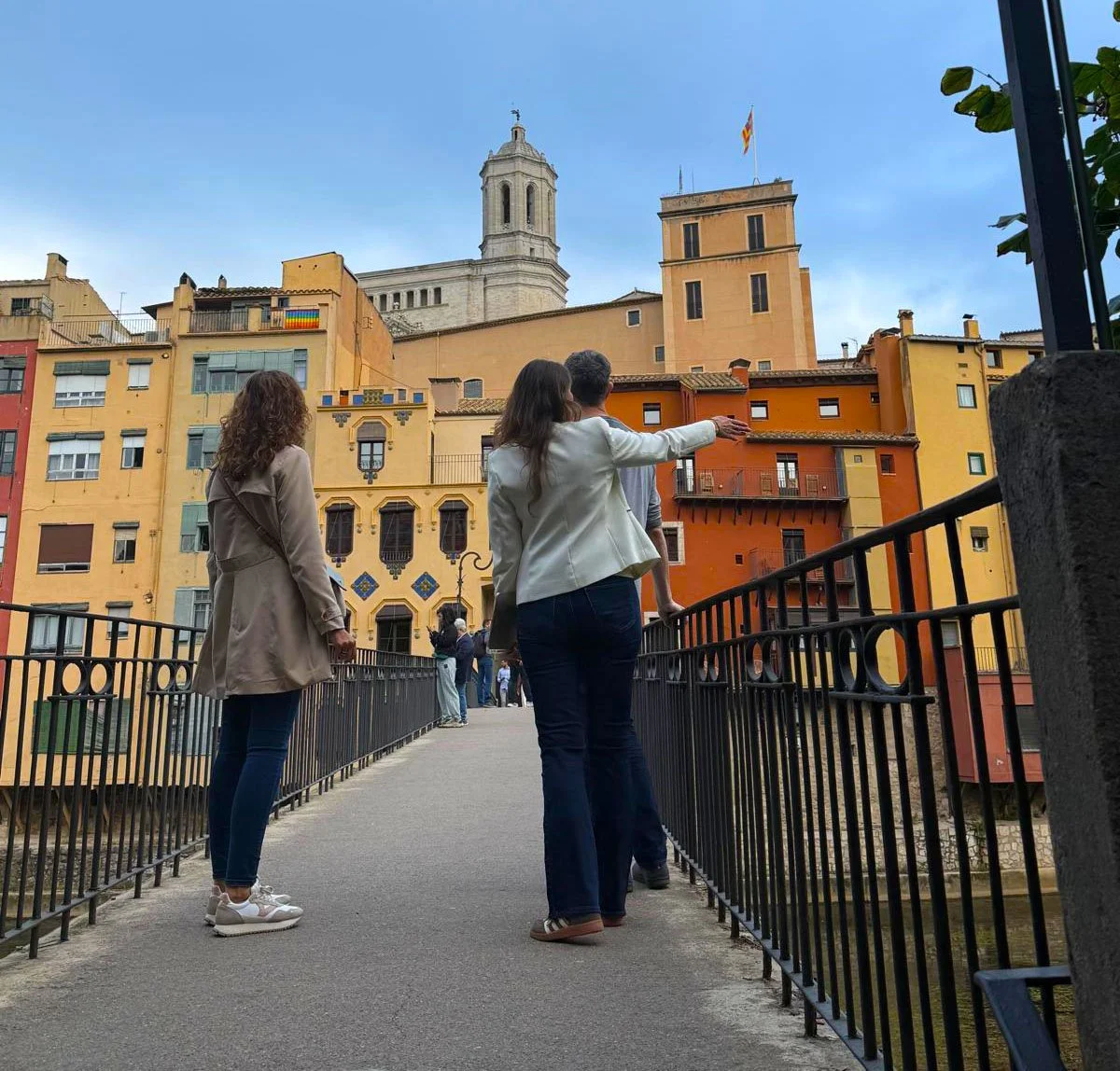 Three women standing on a bridge overlooking colorful old buildings in Barcelona, Spain, with a historic tower in the background.