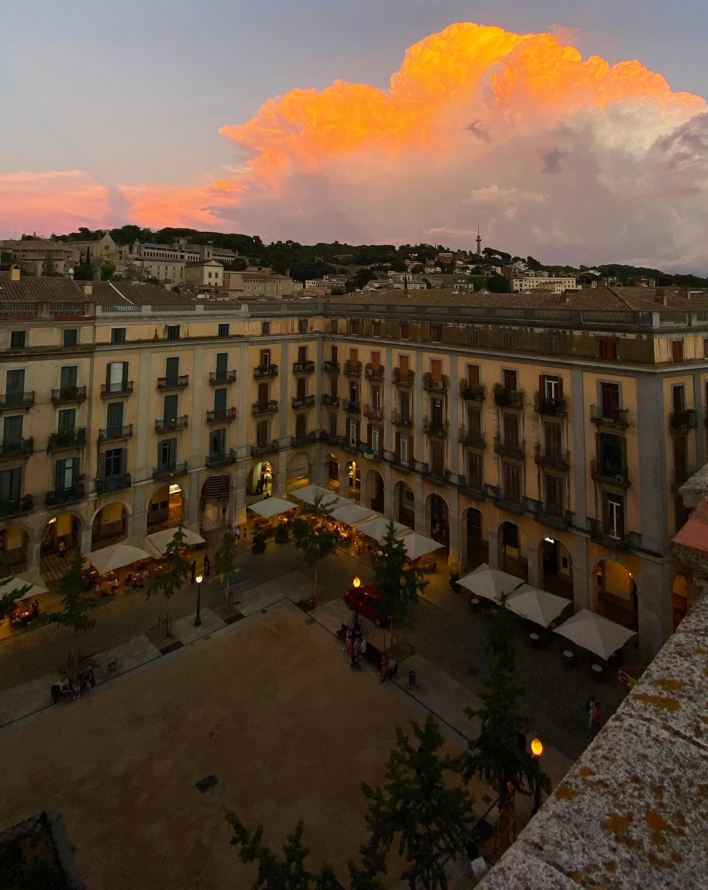 View of a city square at sunset with buildings, streetlights, outdoor dining areas, trees, and a dramatic cloud formation in the sky.