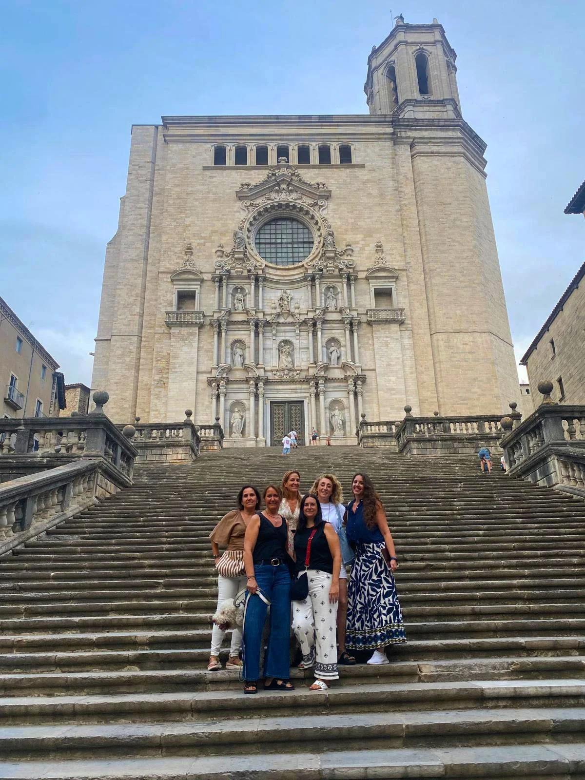 Group of five women standing on stairs in front of a historic church with ornate facade and tall bell tower in Europe.
