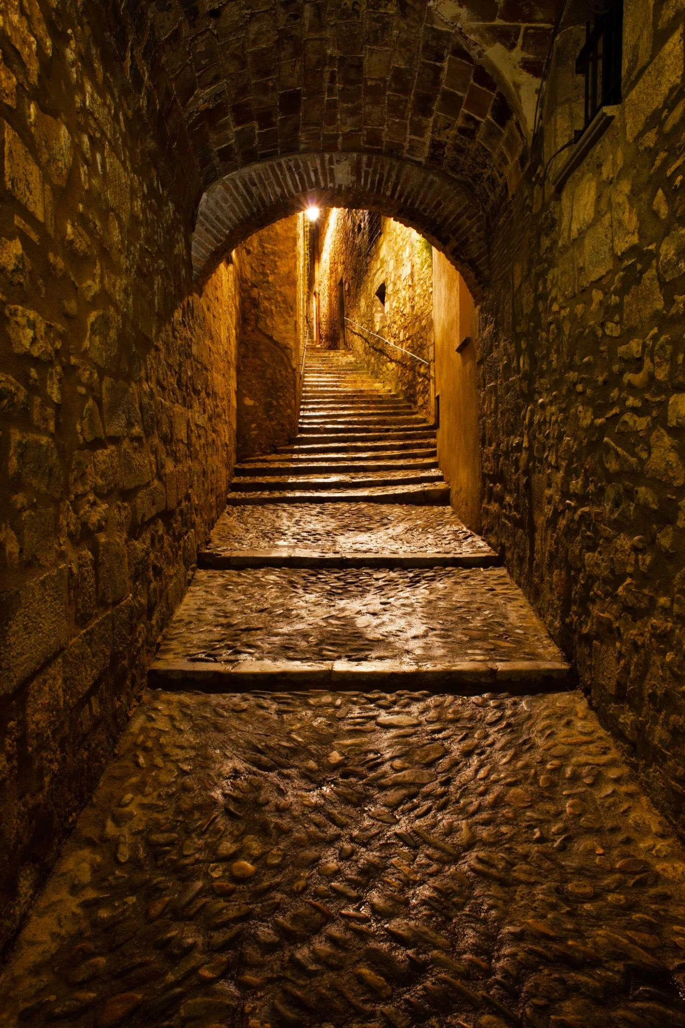 A narrow, cobblestone alleyway with stone stairs leading upward, illuminated by warm yellow-orange lighting under a stone arch.