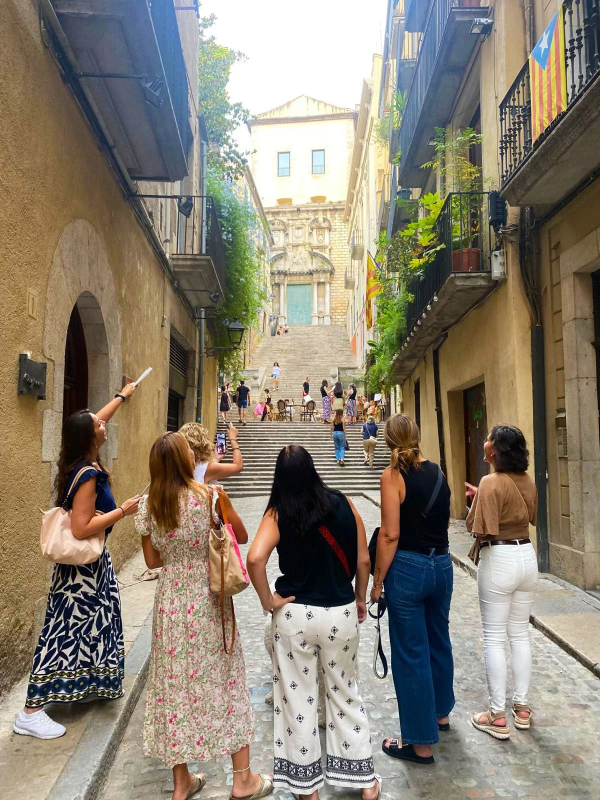 Tourists standing on a cobblestone street in a European city, with stairs leading up to historical buildings and a church in the background, some taking photos, with Catalan flags hanging from buildings.