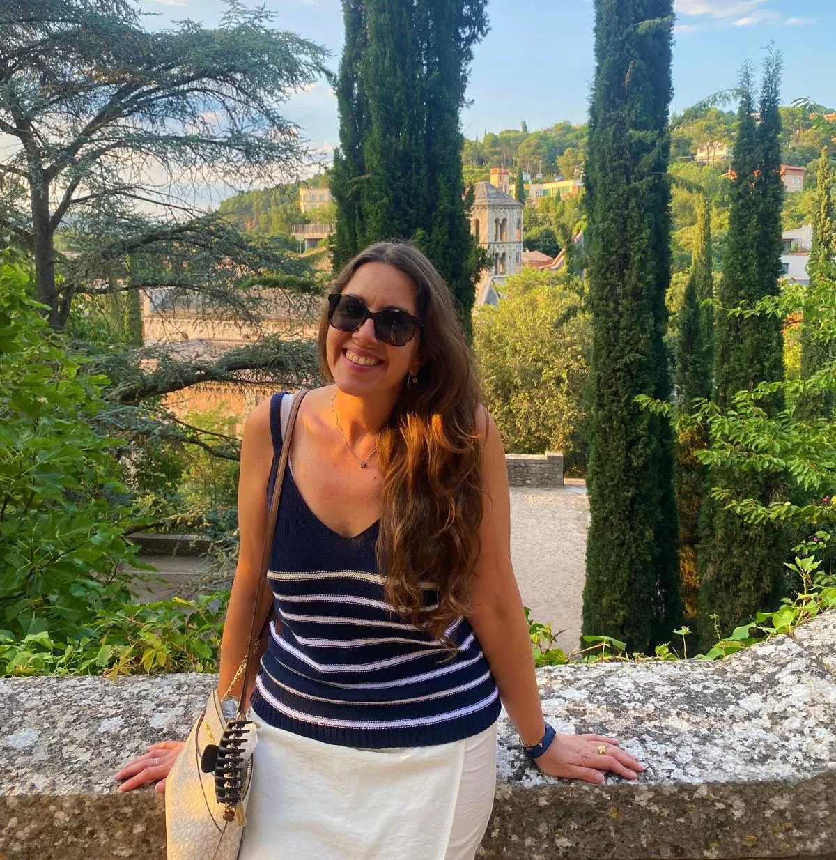 A woman with long wavy hair, sunglasses, and a navy blue striped top, smiling and leaning on a stone wall outdoors with lush green trees and hills in the background.