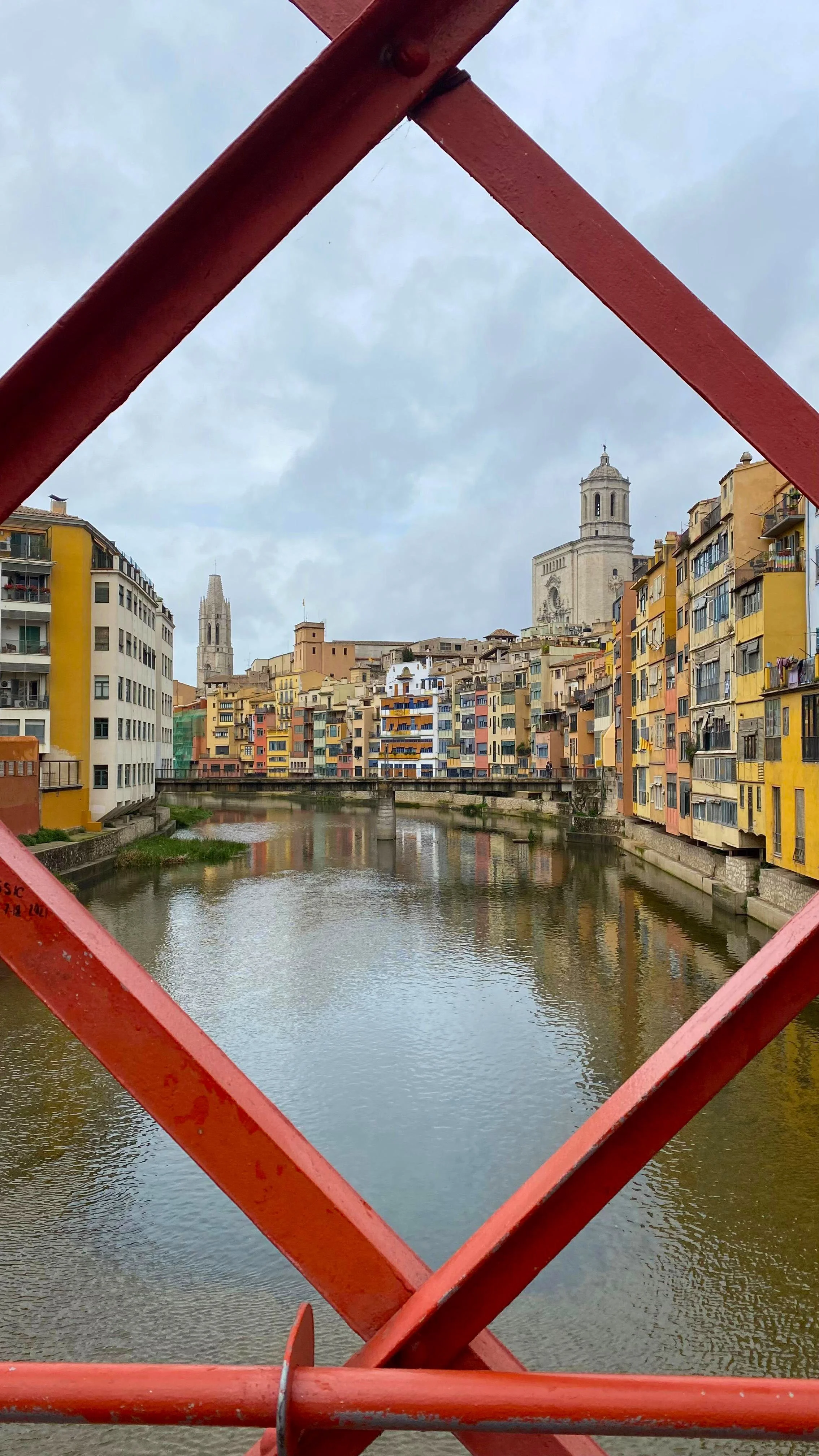 Colorful buildings along a river, viewed through a red metal bridge structure, with a cloudy sky overhead.