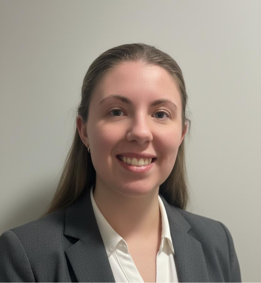 Portrait of a female doctor with long brown hair wearing a dark grey blazer and white shirt, smiling, against a plain light-coloured wall.