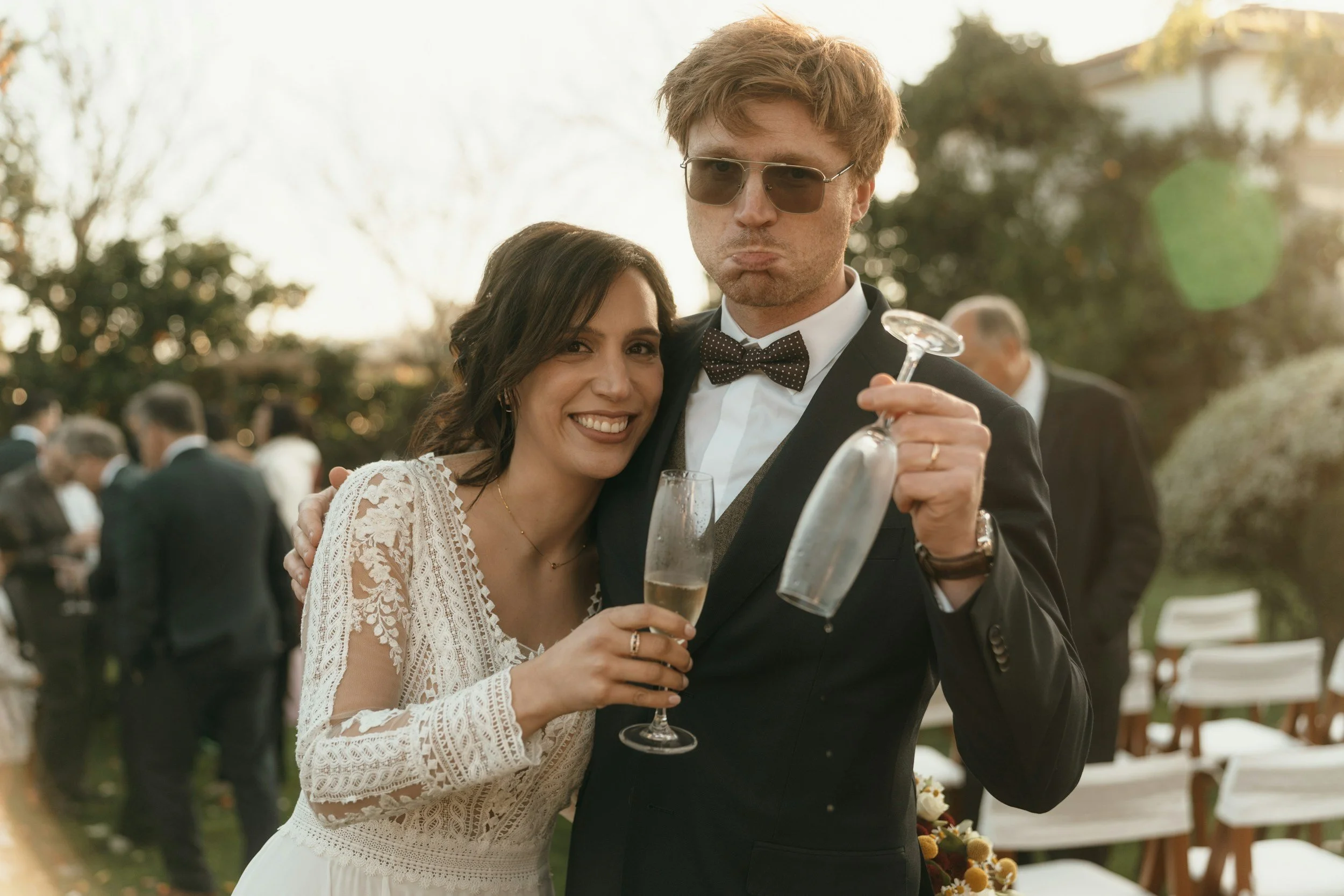 Glückliches Brautpaar beim Anstoßen: Braut im Spitzenkleid lächelt, Bräutigam mit Sonnenbrille und Fliege zieht eine lustige Grimasse mit Sektglas.