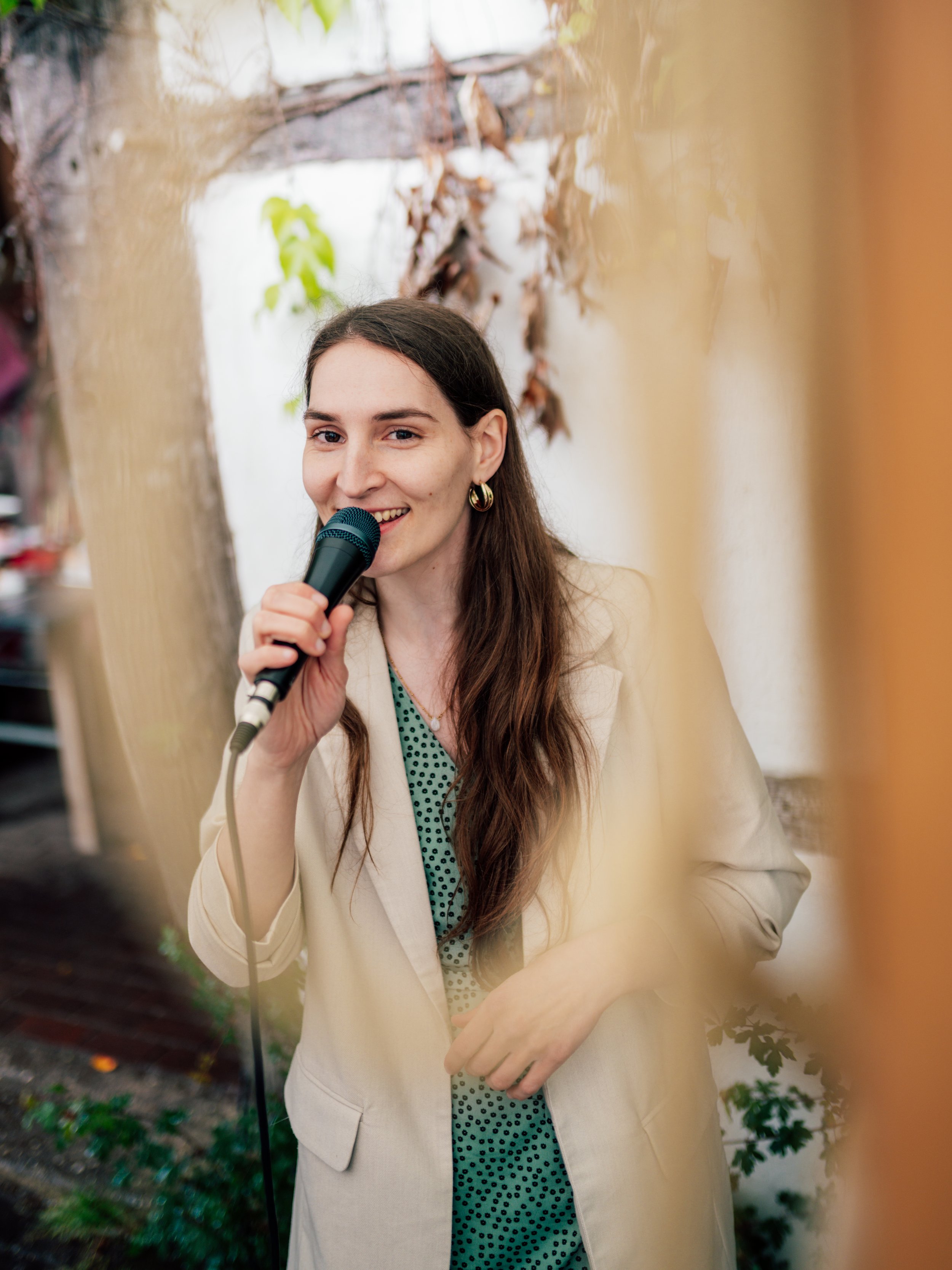 Eine junge Frau mit langen braunen Haaren, die eine Hand in der Tasche hat, hält ein Mikrofon und lächelt in die Kamera, während sie spricht, im Hintergrund sind Blätter und eine Wand zu sehen.