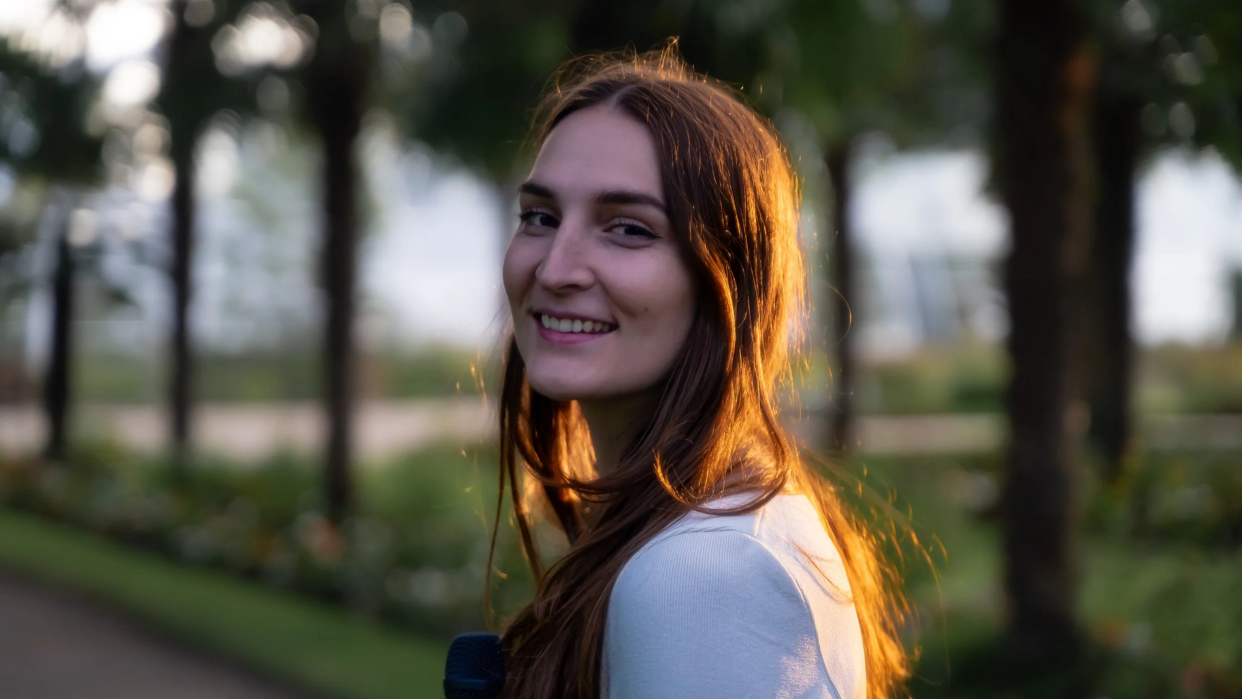 Junge Frau mit langen roten Haaren, die in einem Park bei Sonnenuntergang lächelt.