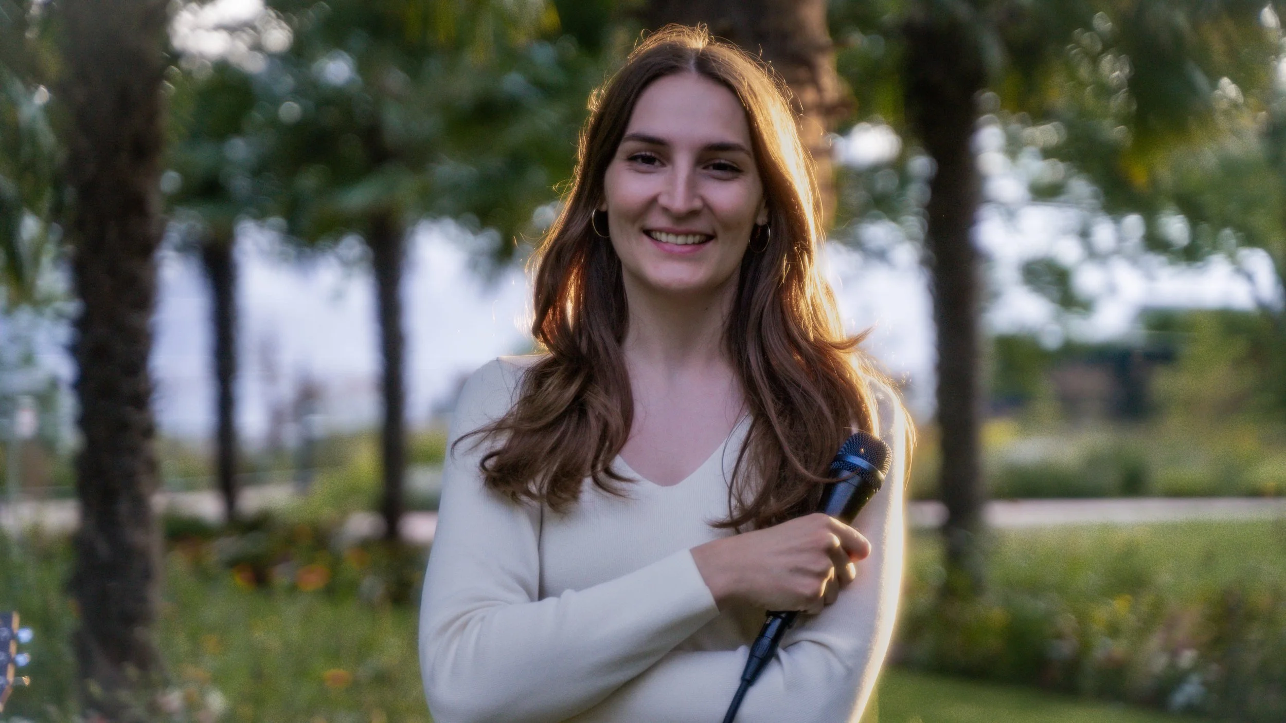 Lächelnde junge Frau mit einem Mikrofon im Freien, umgeben von Bäumen und Grünflächen.