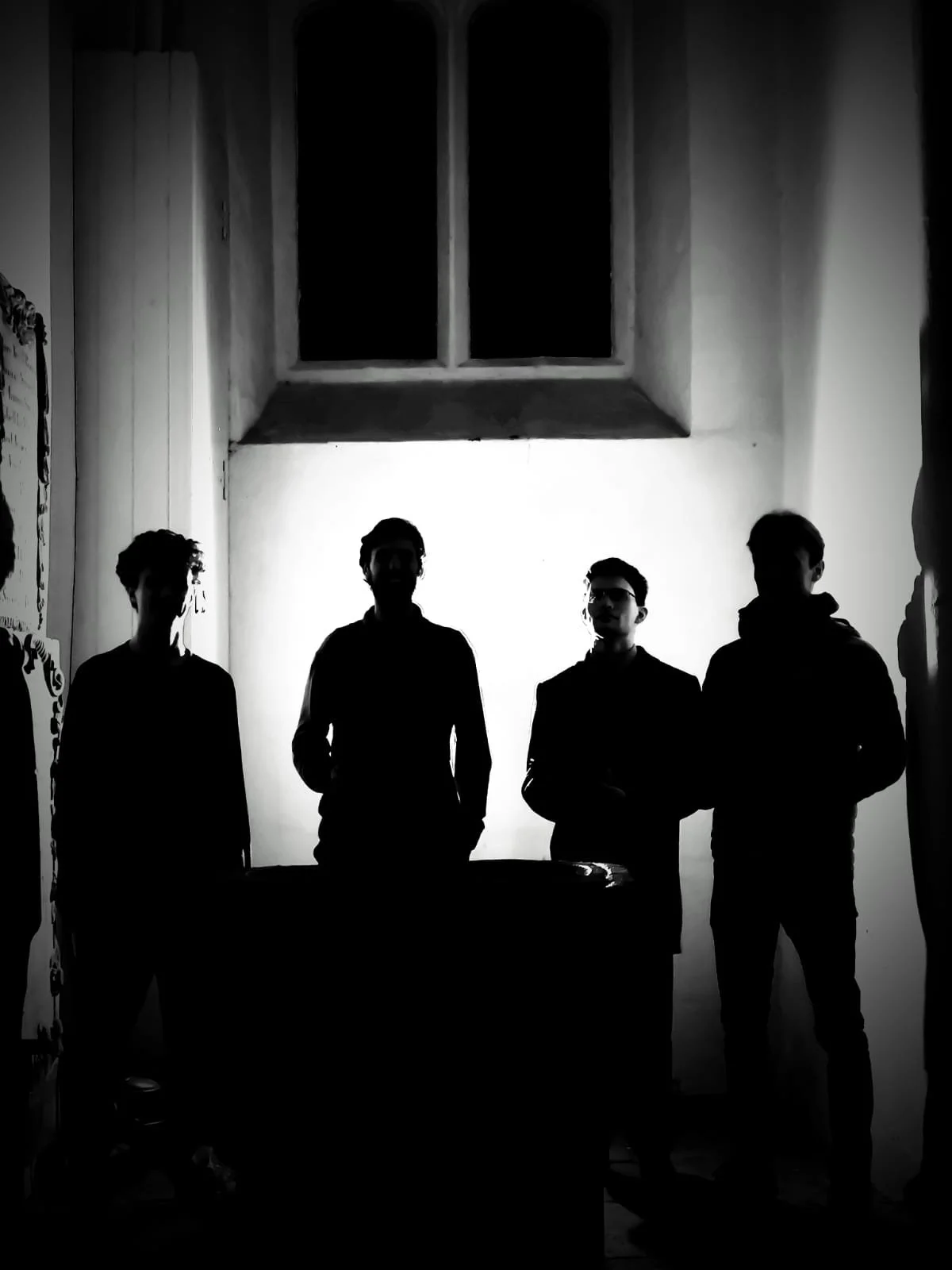 Four silhouettes of young men standing in a dimly lit room with a window overhead, creating a dramatic shadow effect.