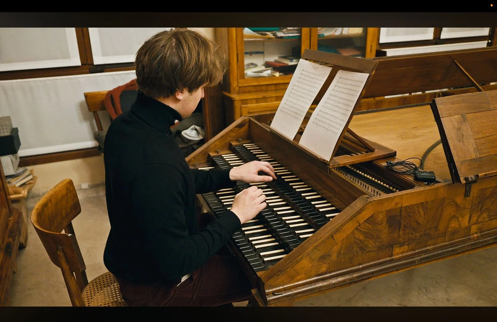 A young man with brown hair, wearing a black turtleneck, plays a wooden upright piano in a room with bookshelves and filing cabinets.