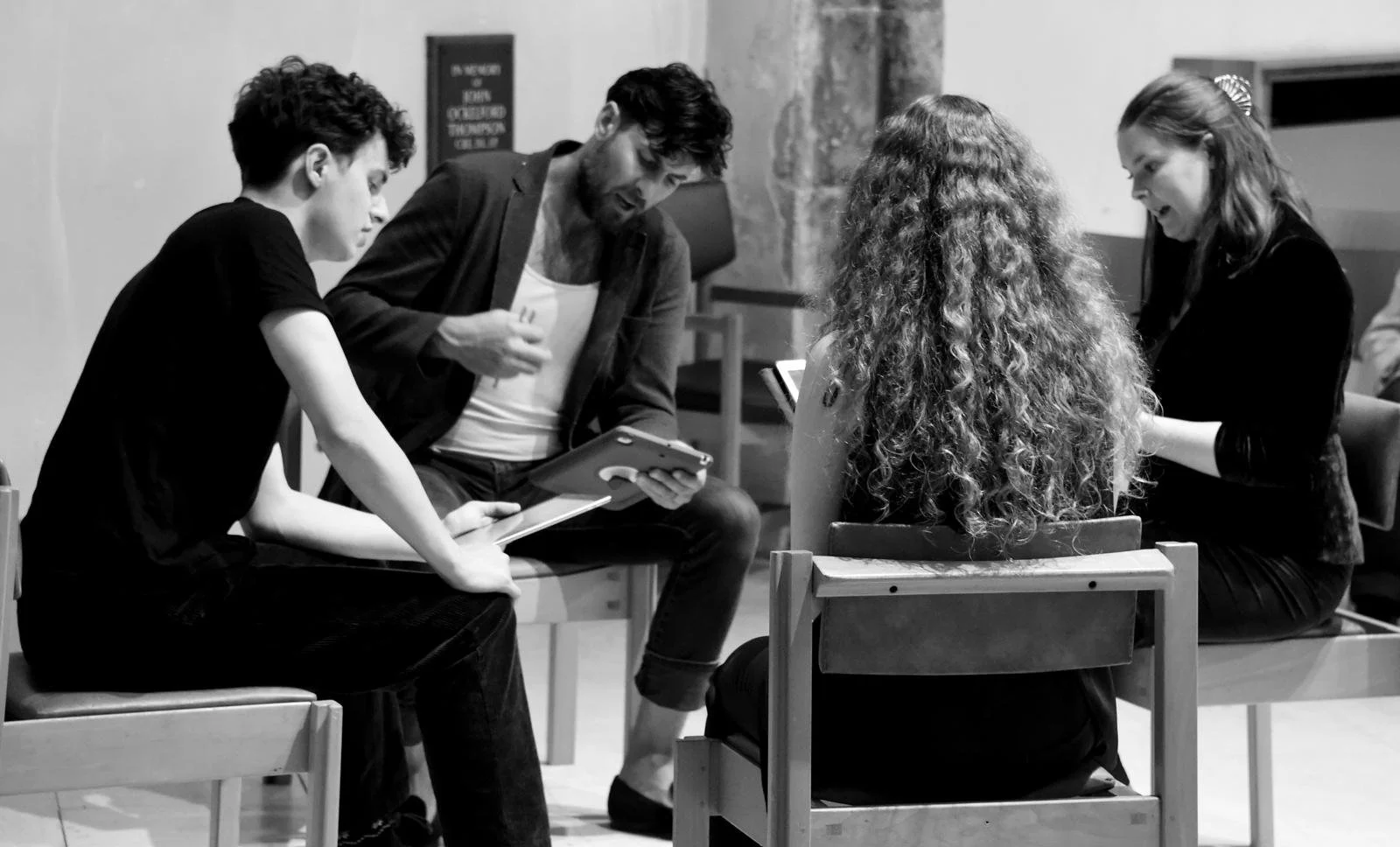 Group of four people sitting in a circle, engaged in conversation and looking at tablets and phones, in black and white.