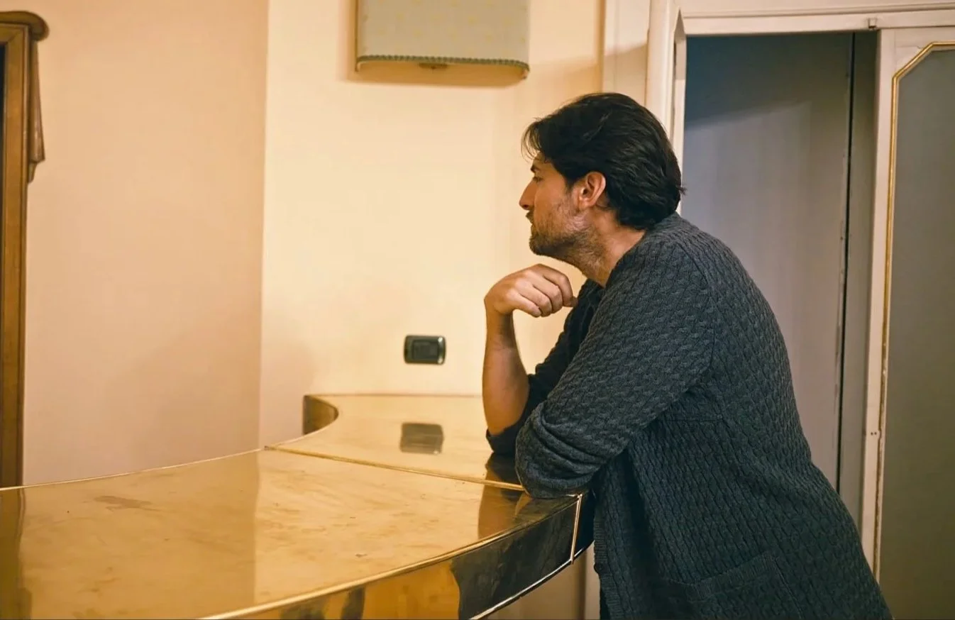 A man with dark hair and beard, wearing a dark sweater, leaning on a wooden piano, looking to the right in a room with beige walls and a mirror.