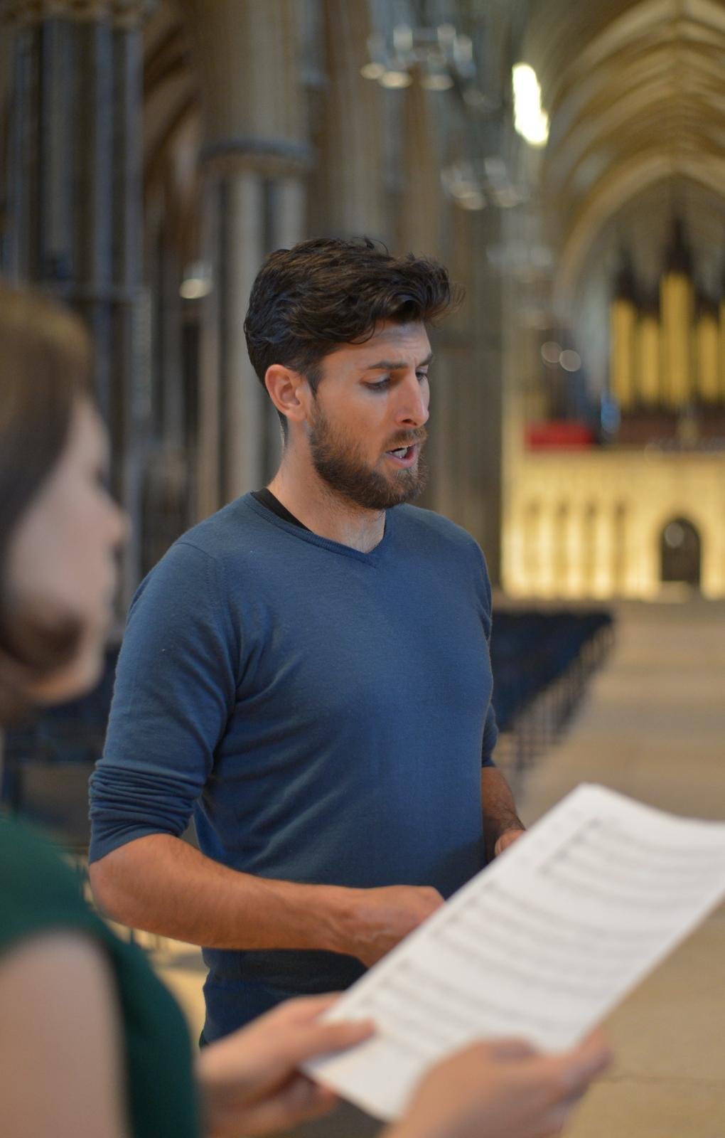 A man with dark hair and a beard, wearing a blue shirt, appears to be singing or speaking in a church or cathedral, with stained glass windows and an altar in the background. A woman is partially visible in the foreground, holding a sheet of music or a program.