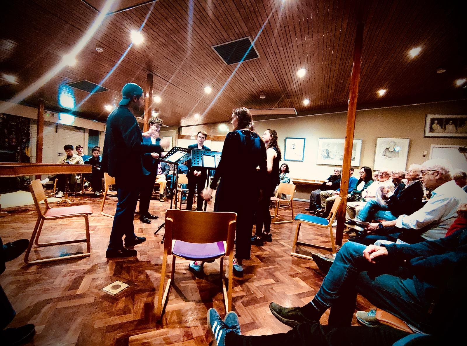 Group of people performing music in a cozy, wood-paneled room with an audience watching.