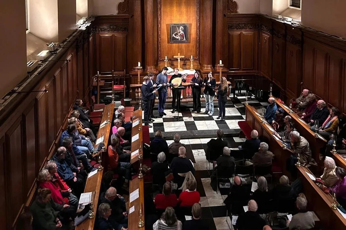A musical performance takes place in a church or chapel, with a group of six musicians playing instruments on a platform in front of an audience seated on benches and chairs. The background features wooden paneling, a small cross, and a framed portrait.