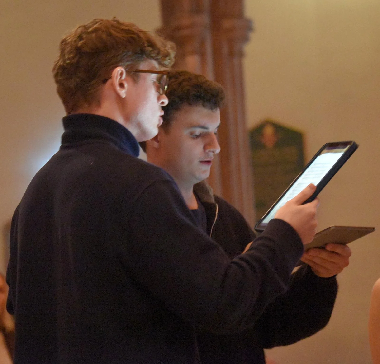 Two young men stand indoors, looking at their tablets or phones, in a setting that appears to be a church or historic building.
