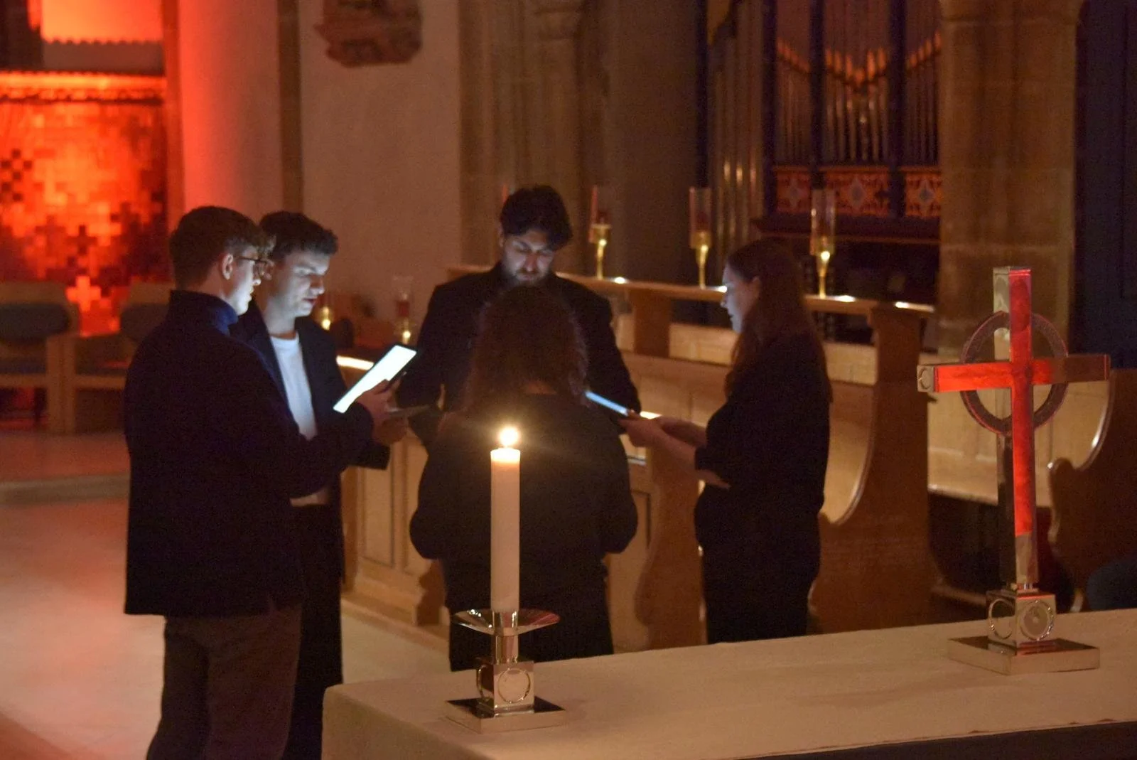 Five people standing around a table inside a dimly lit church, several are looking at their phones, a lit candle on the table, and a decorative cross on the right.