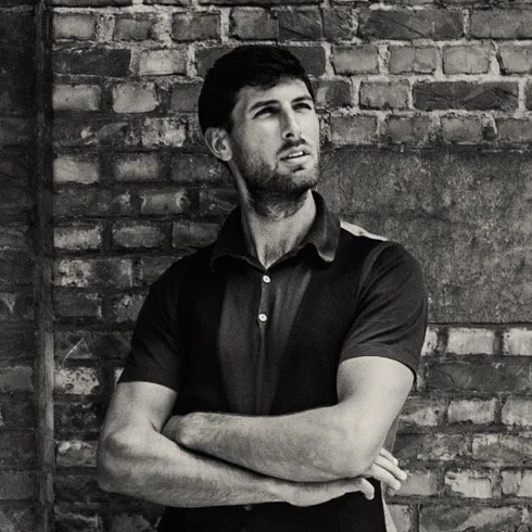A young man with dark hair and a beard standing in front of a brick wall, looking to the side with arms crossed.