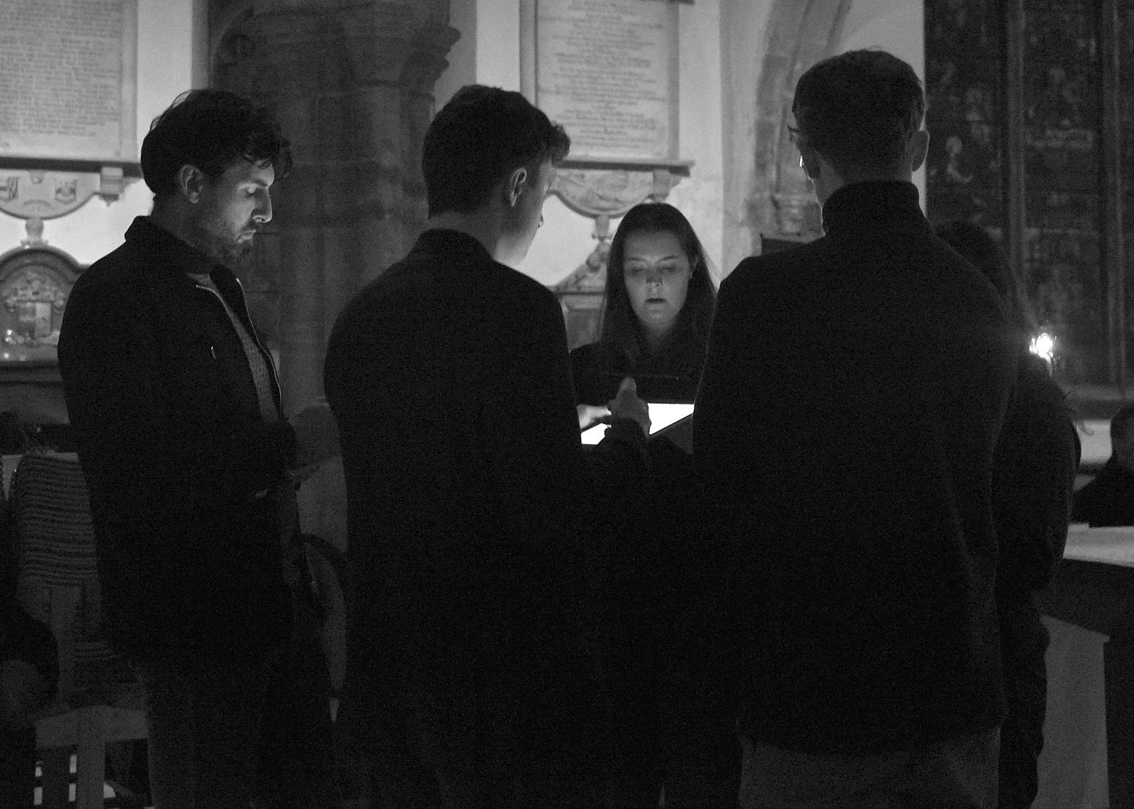Four people standing around a table looking at a tablet in a dimly lit room with historical paintings on the walls.
