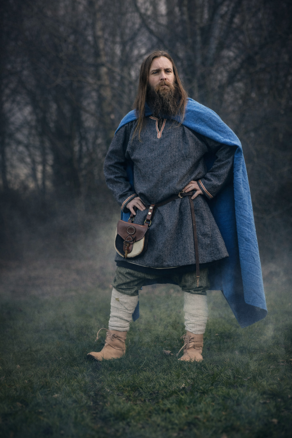 A man dressed in medieval attire standing outdoors in a field with trees in the background. He has long hair and a beard, wearing a dark tunic, a blue cloak, and medieval-style boots and leg wraps, with his hands on his hips.