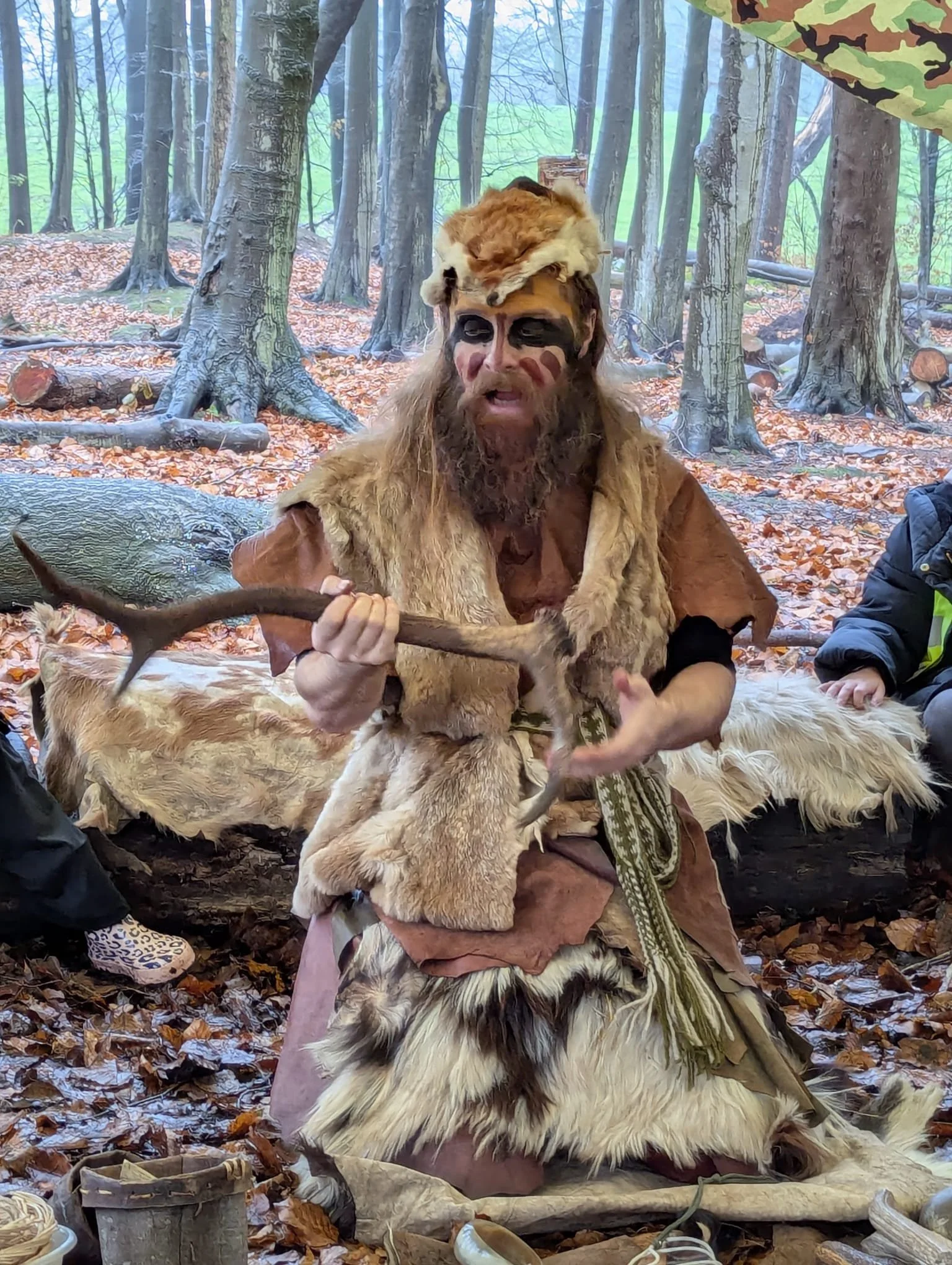 A person dressed as a Stone Age or primitive tribal character sitting in a forest, holding a spear, with face paint, a fur and animal hide costume, and a headdress made of an animal's skull and fur. The background is a wooded area with trees and fallen autumn leaves on the ground.