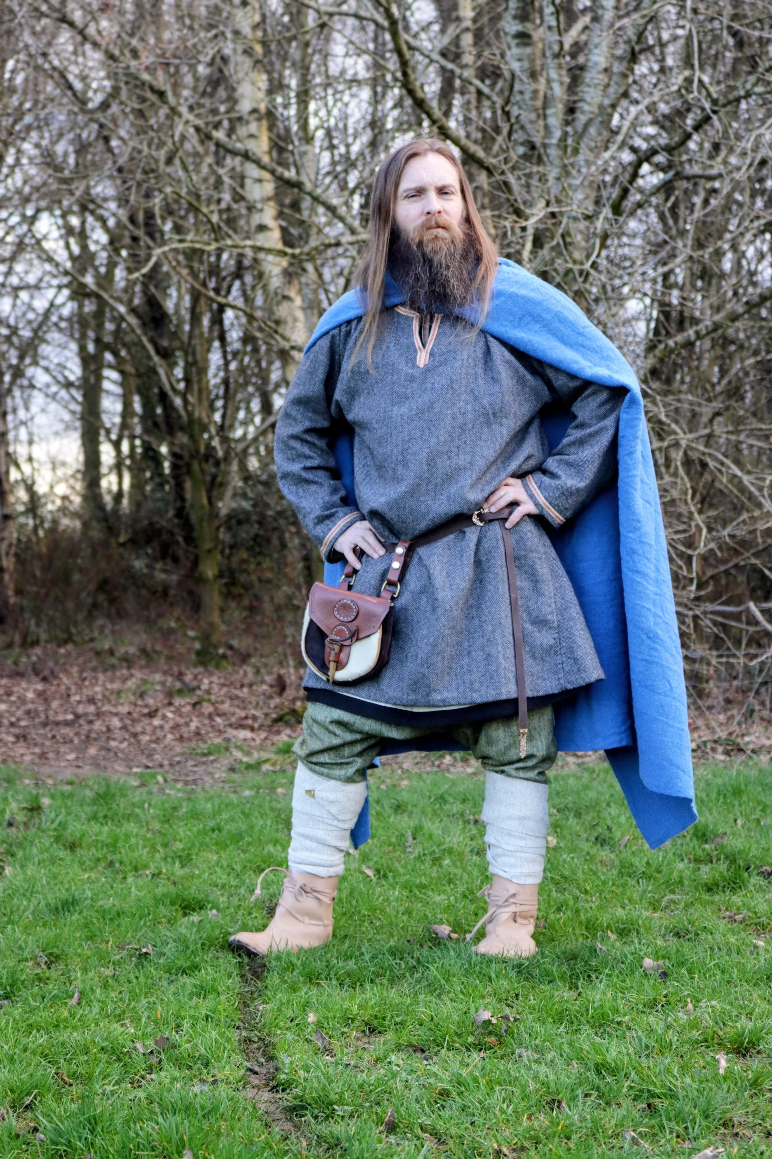 A man dressed as a medieval or fantasy character stands on a grassy lawn with trees in the background. He has long hair and a beard, wearing a gray tunic, a blue cloak, green pants, and beige boots with white leg wraps. He has one hand on his hip and appears to be posing confidently.