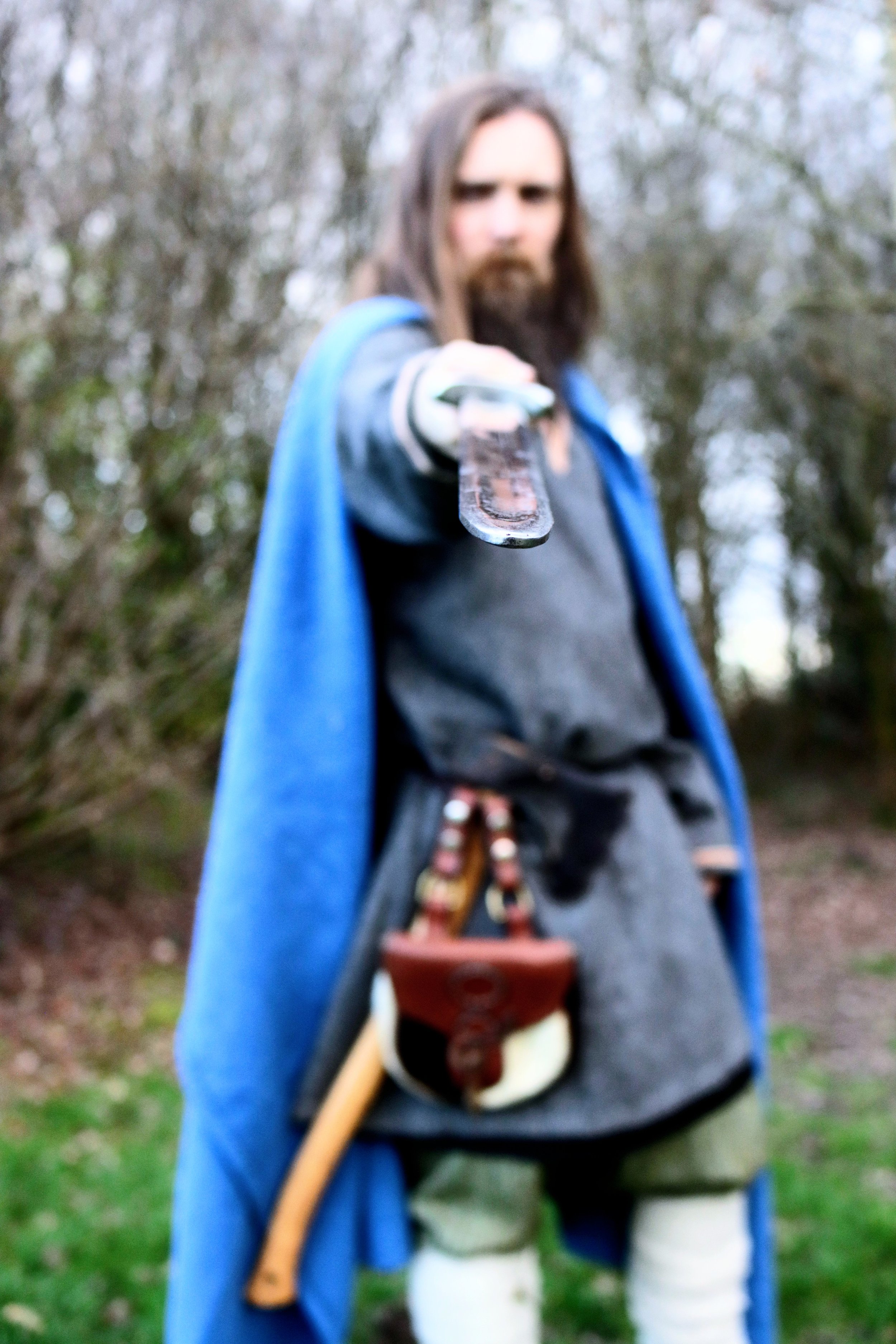 A man dressed as a fantasy or medieval character holds a sword towards the camera in an outdoor setting with trees.
