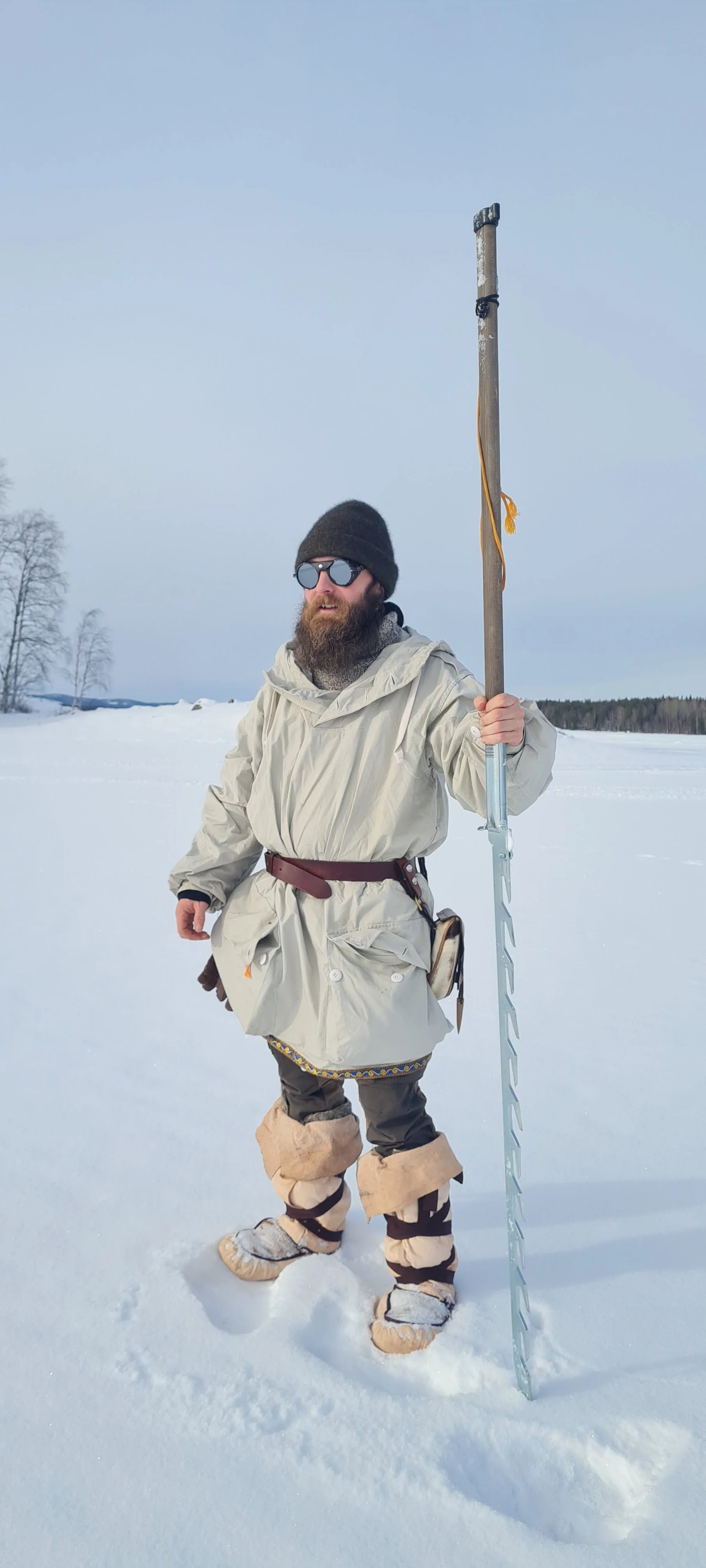 A man with a beard dressed in traditional winter clothing holding a wooden staff in a snowy landscape.