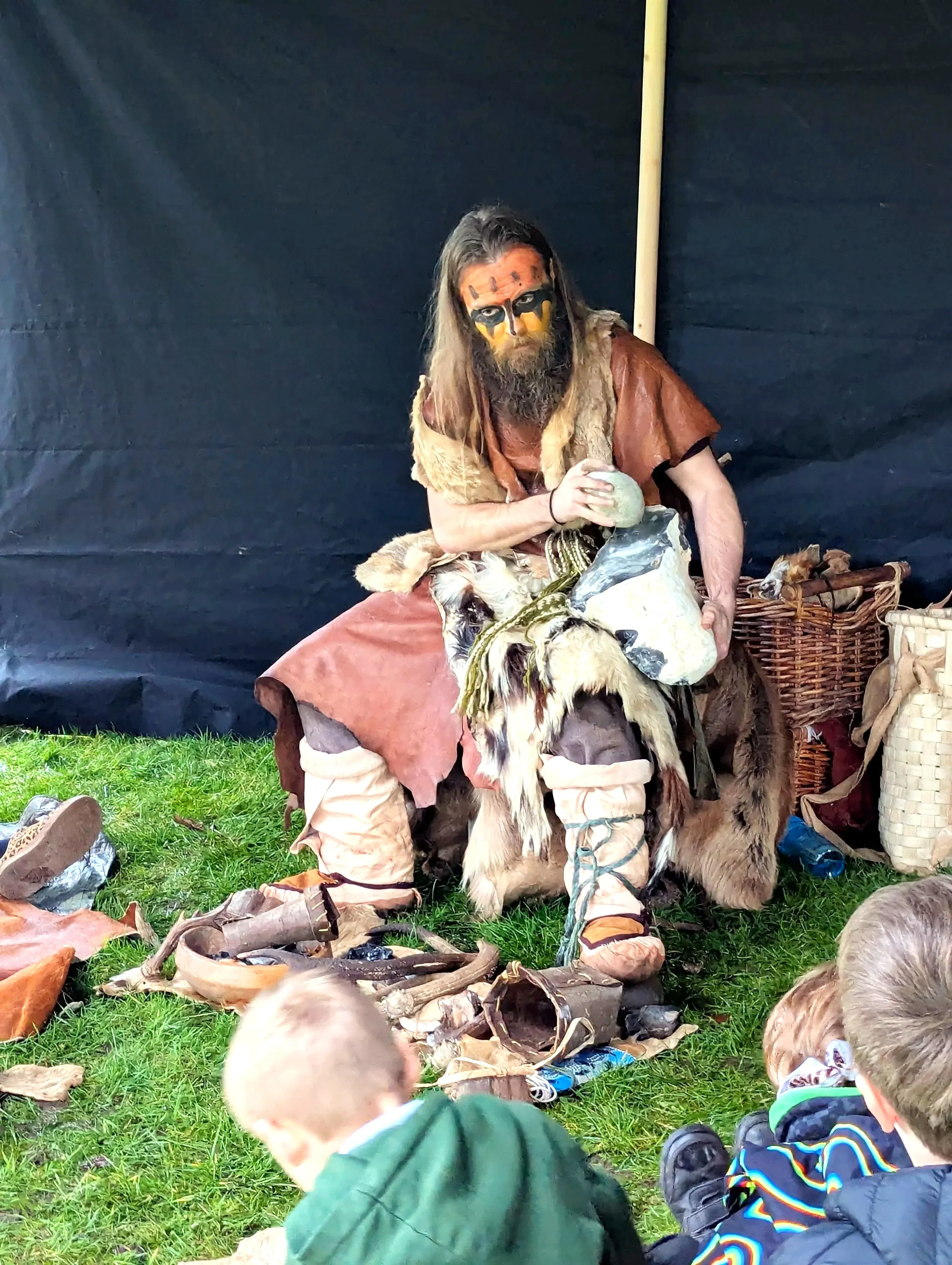A man dressed as a prehistoric caveman with face paint, long hair, and a beard, sitting on a chair or prop designed to look like a rock, surrounded by props and costumes resembling primitive tools and animals, with children watching in the foreground.