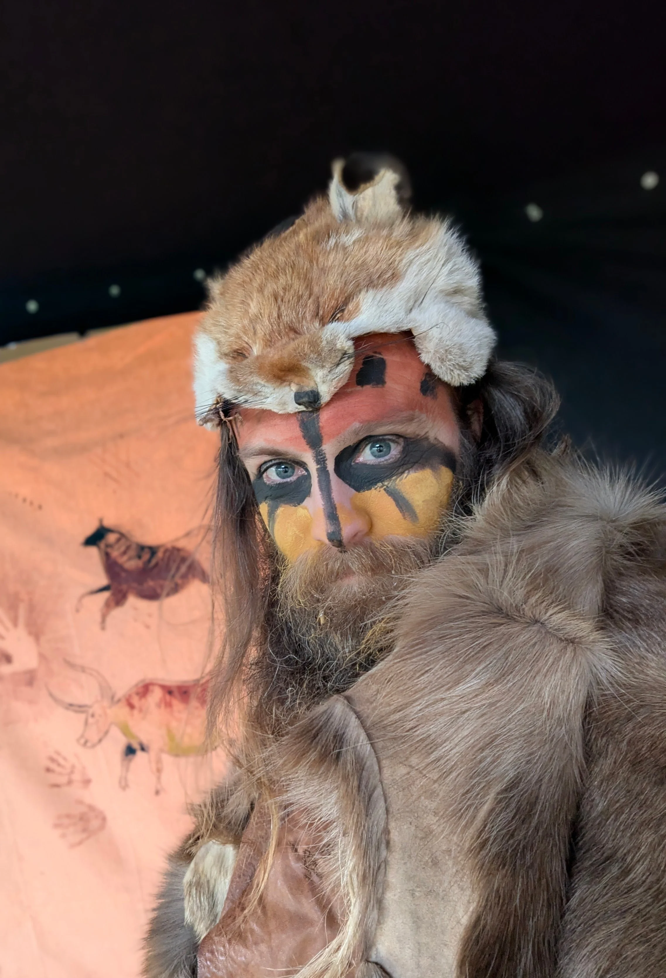 A person with face paint resembling a bee, wearing animal fur clothing, and a dead fox draped over their head. In the background, there is a painting of various animals, including a buffalo and an elk.