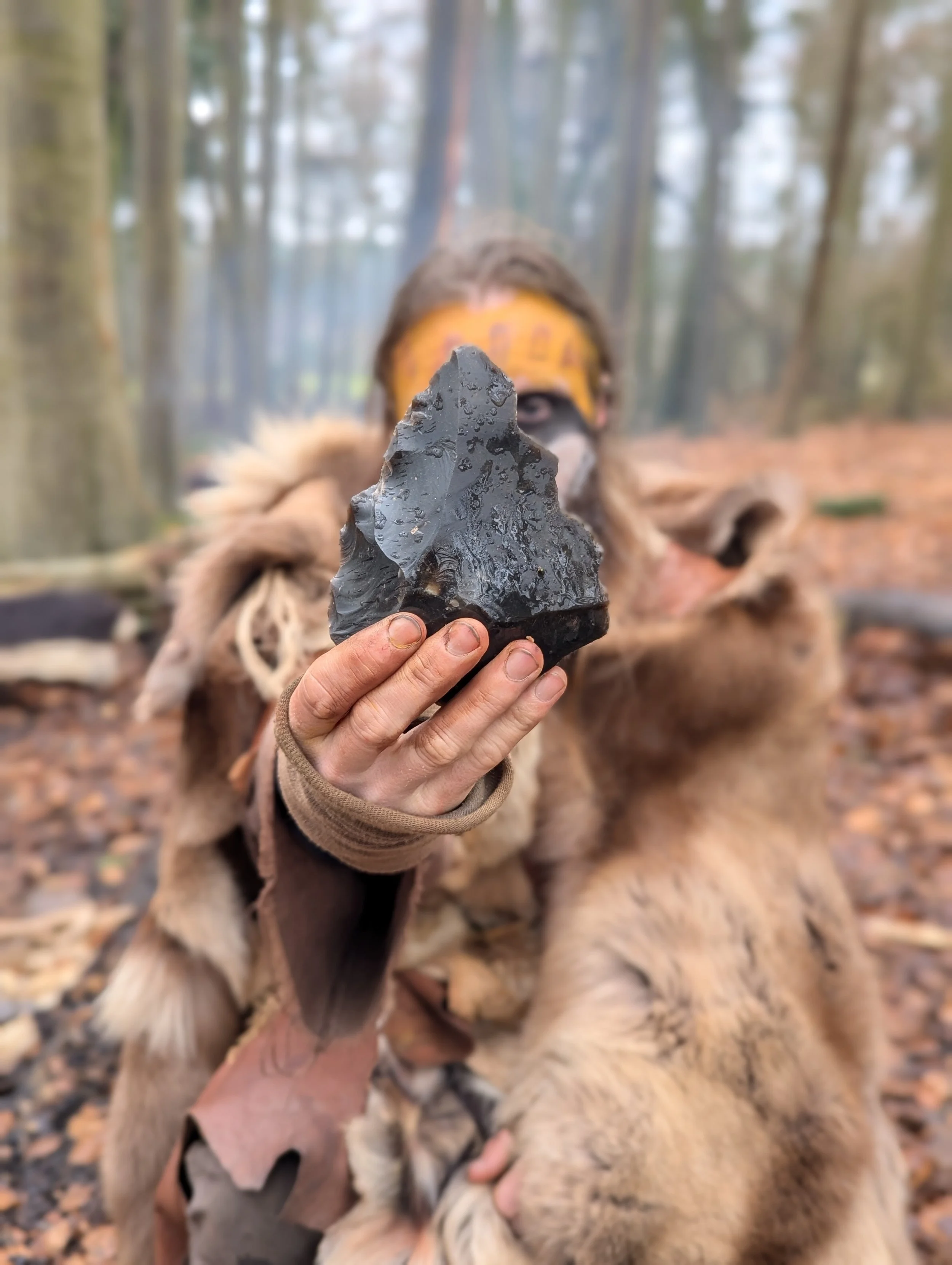 A person in prehistoric clothing holding a large, sharp arrowhead in front of their face in a forest setting.