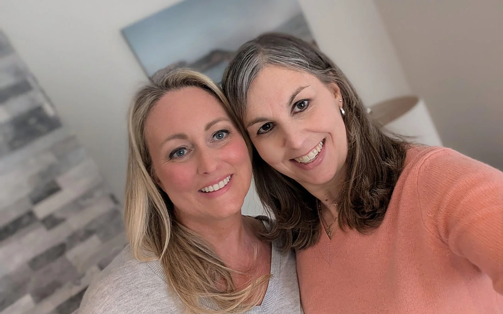 Two women smiling for a selfie indoors, with a modern stone wall and a lamp in the background.