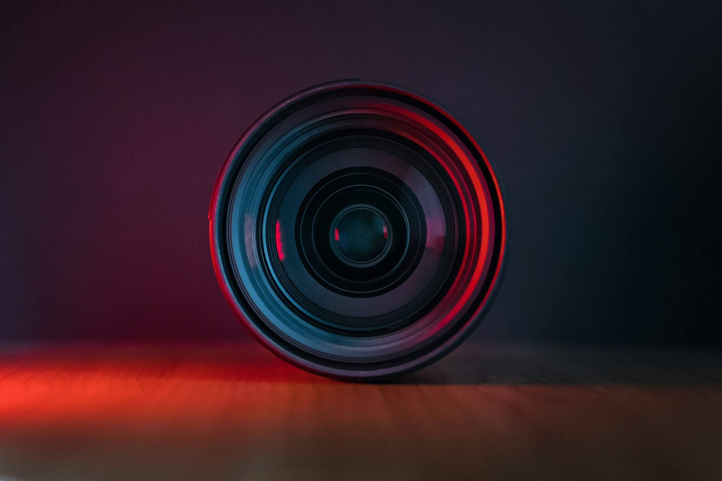 Close-up of a camera lens with red and blue neon lights reflecting on it, against a dark background.