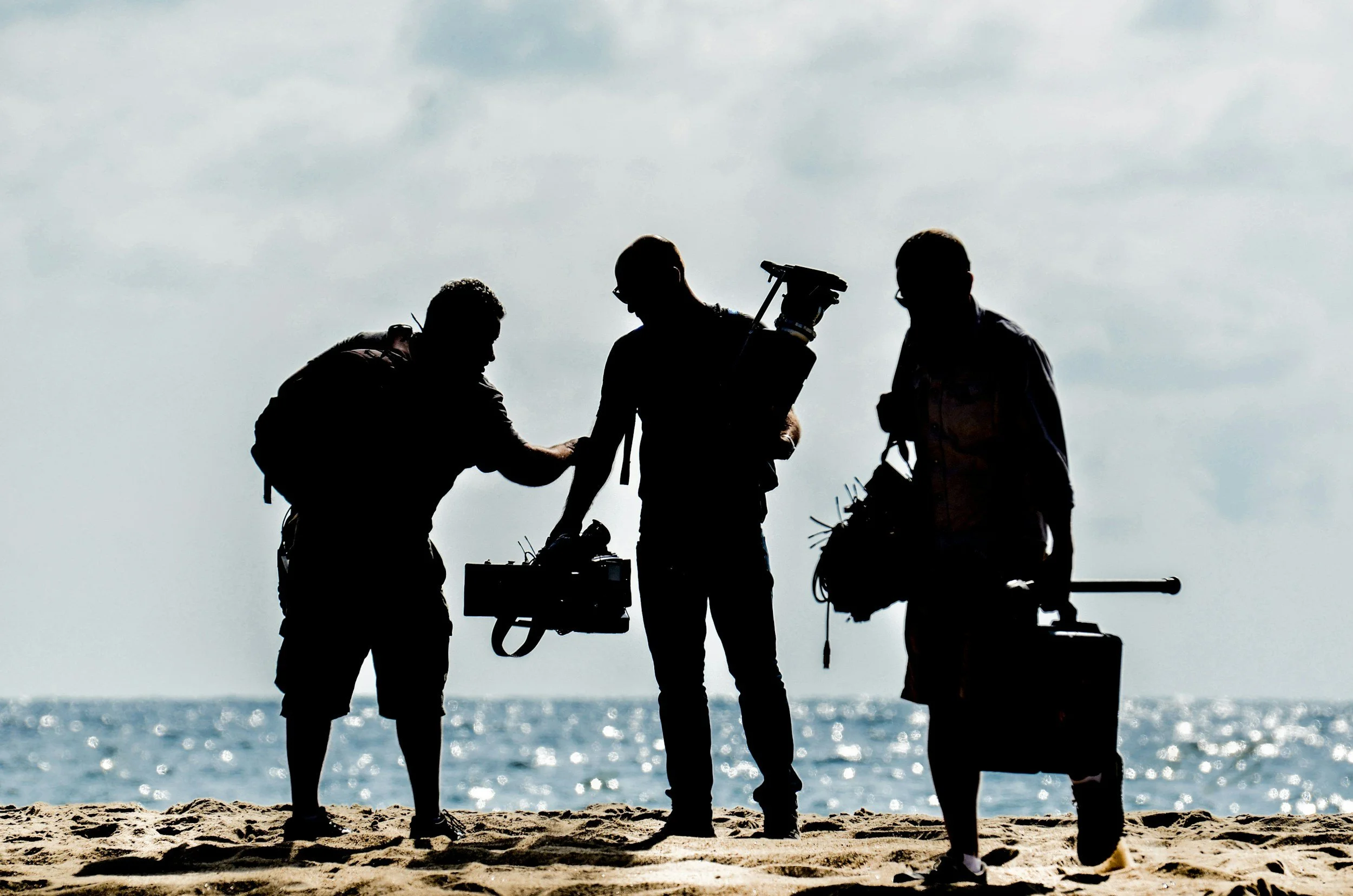 Silhouettes of three people working on a film set on the beach, with the ocean and sky in the background.