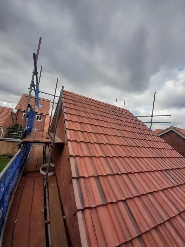 View of a house roof under construction with red roof tiles, scaffolding on the side, and a cloudy sky overhead.
