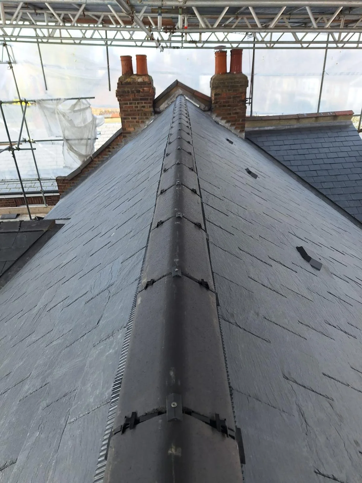 Close-up view of a residential roof with slate shingles and two brick chimneys under construction scaffolding.