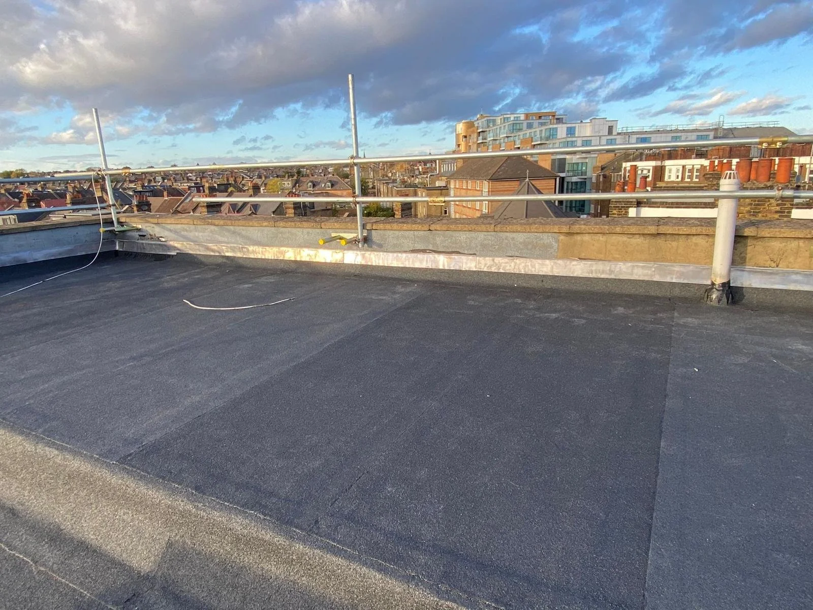 Building rooftop with safety rails, asphalt surface, and a view of city rooftops and modern buildings under a partly cloudy sky.