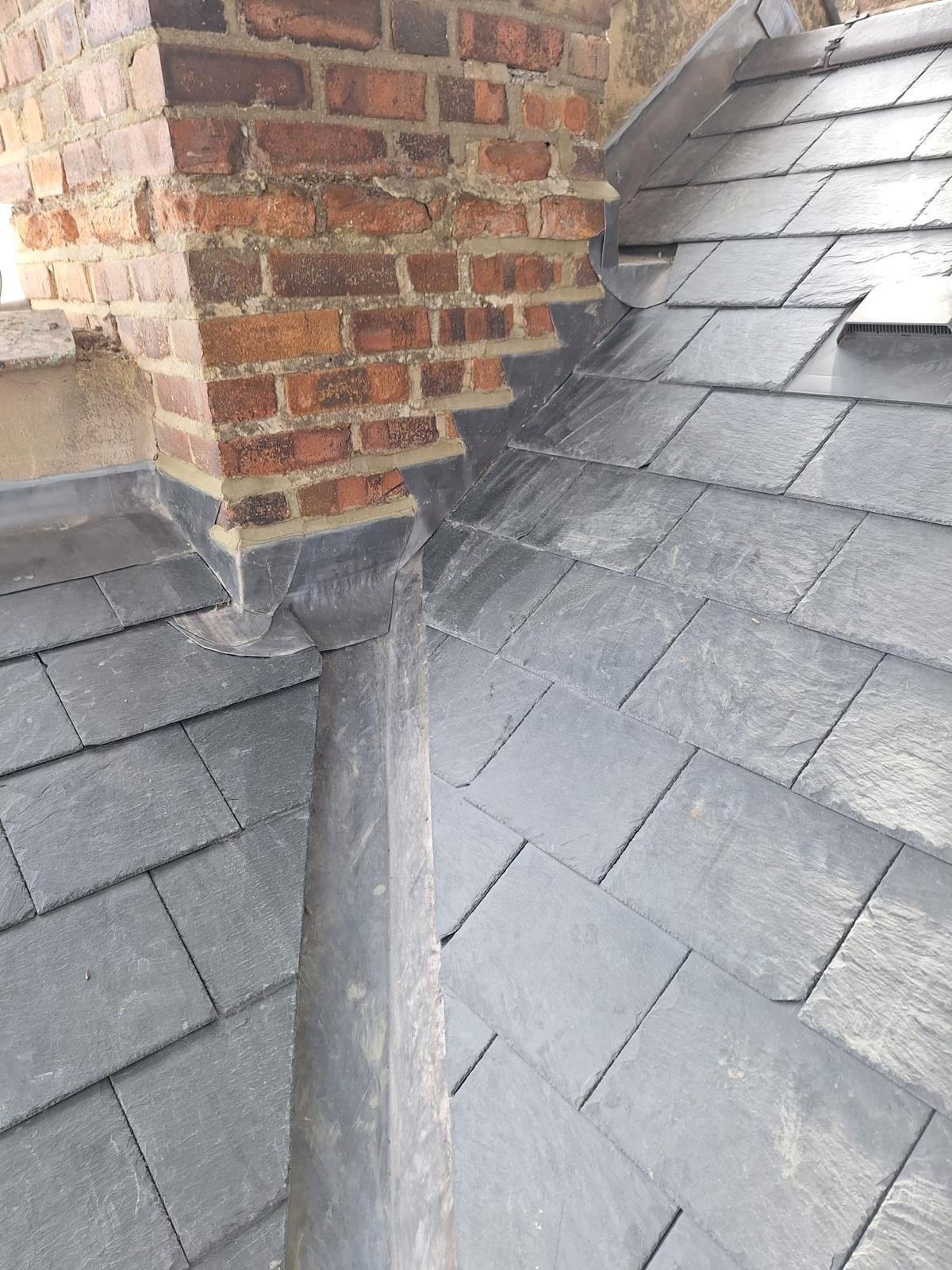 Close-up of a brick chimney with weathered red bricks and mortar next to a slate shingled roof with gray tiles.
