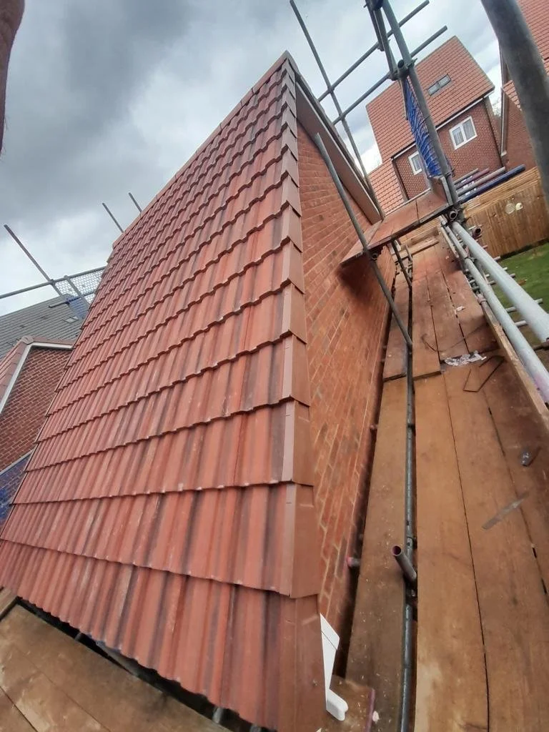 View of a newly installed red tile roof on a brick house from a low angle, with scaffolding around the building under cloudy sky.