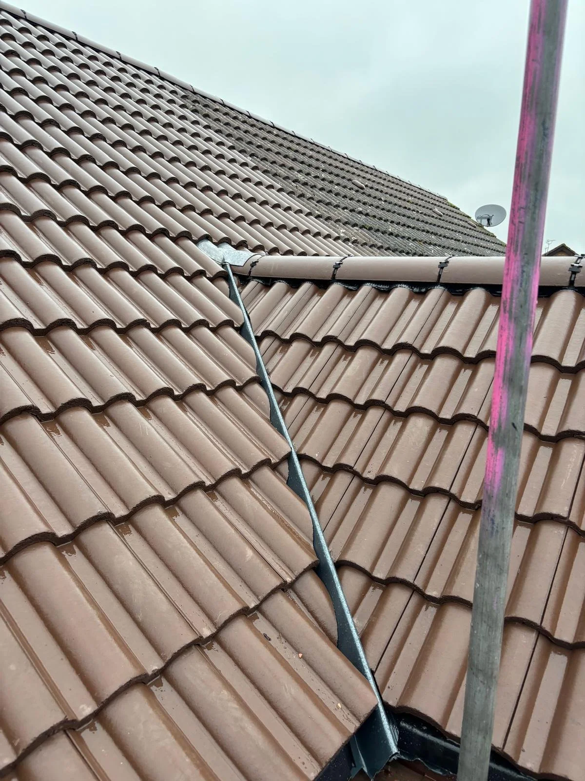 Close-up view of a brown tiled roof with a visible vent pipe and a ladder or scaffolding pole in the foreground, overcast sky in the background.