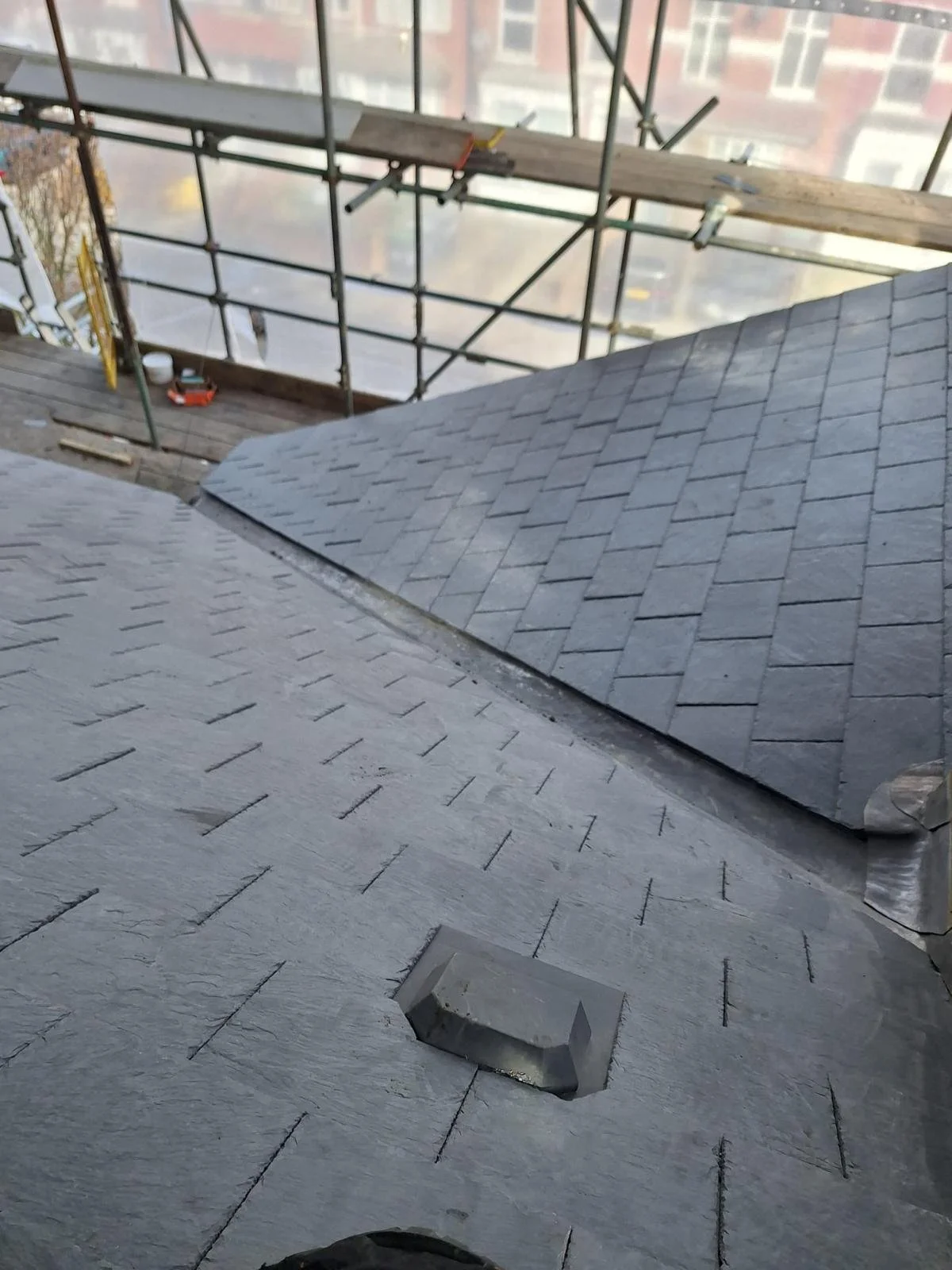 Close-up of a rooftop with black shingles, scaffolding in the background, and building structures visible behind the scaffolding.
