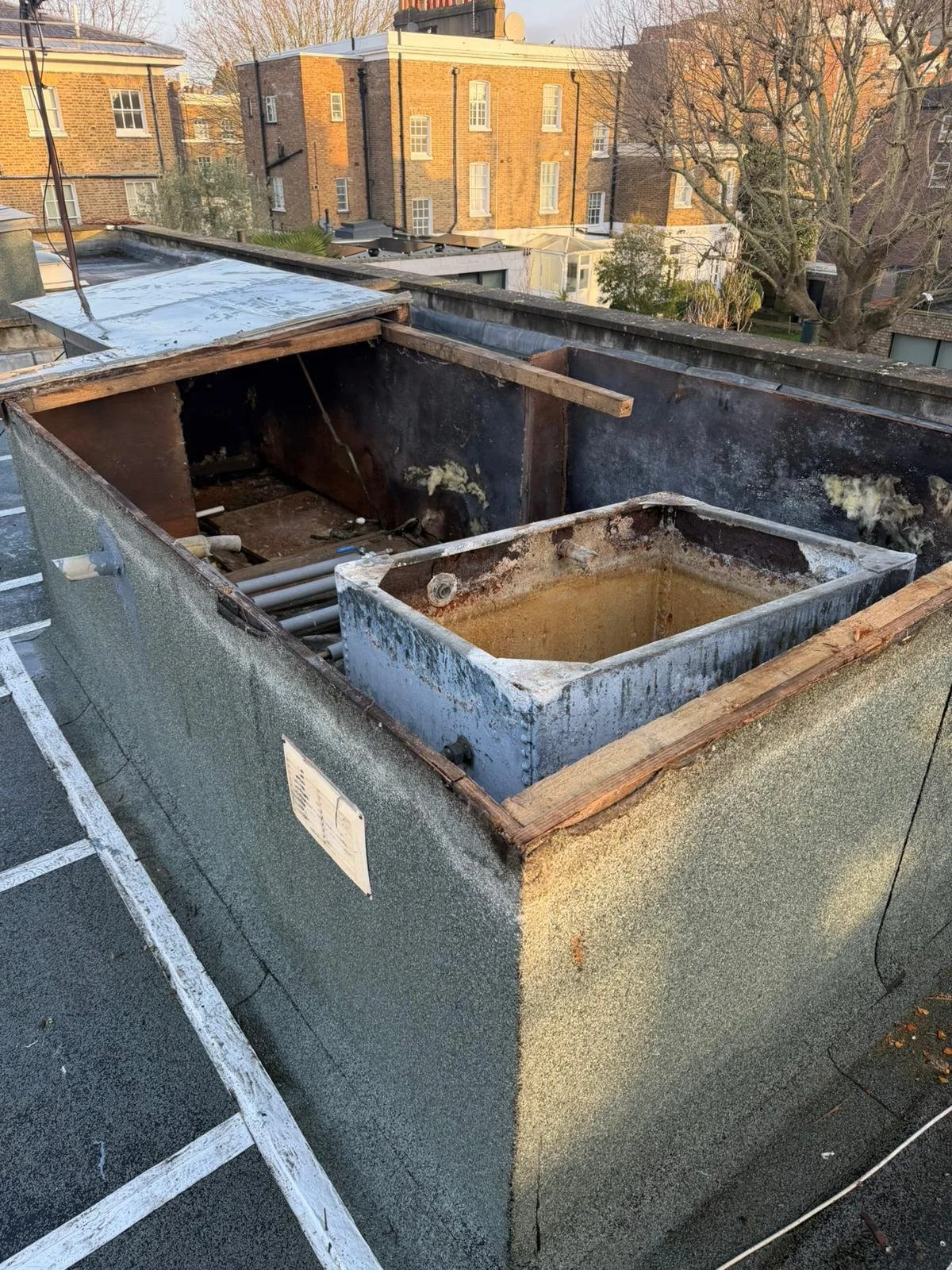 View of a rooftop HVAC unit with its outer cover removed, exposing the internal metal components and pipes, in a city setting with brick buildings and trees in the background.