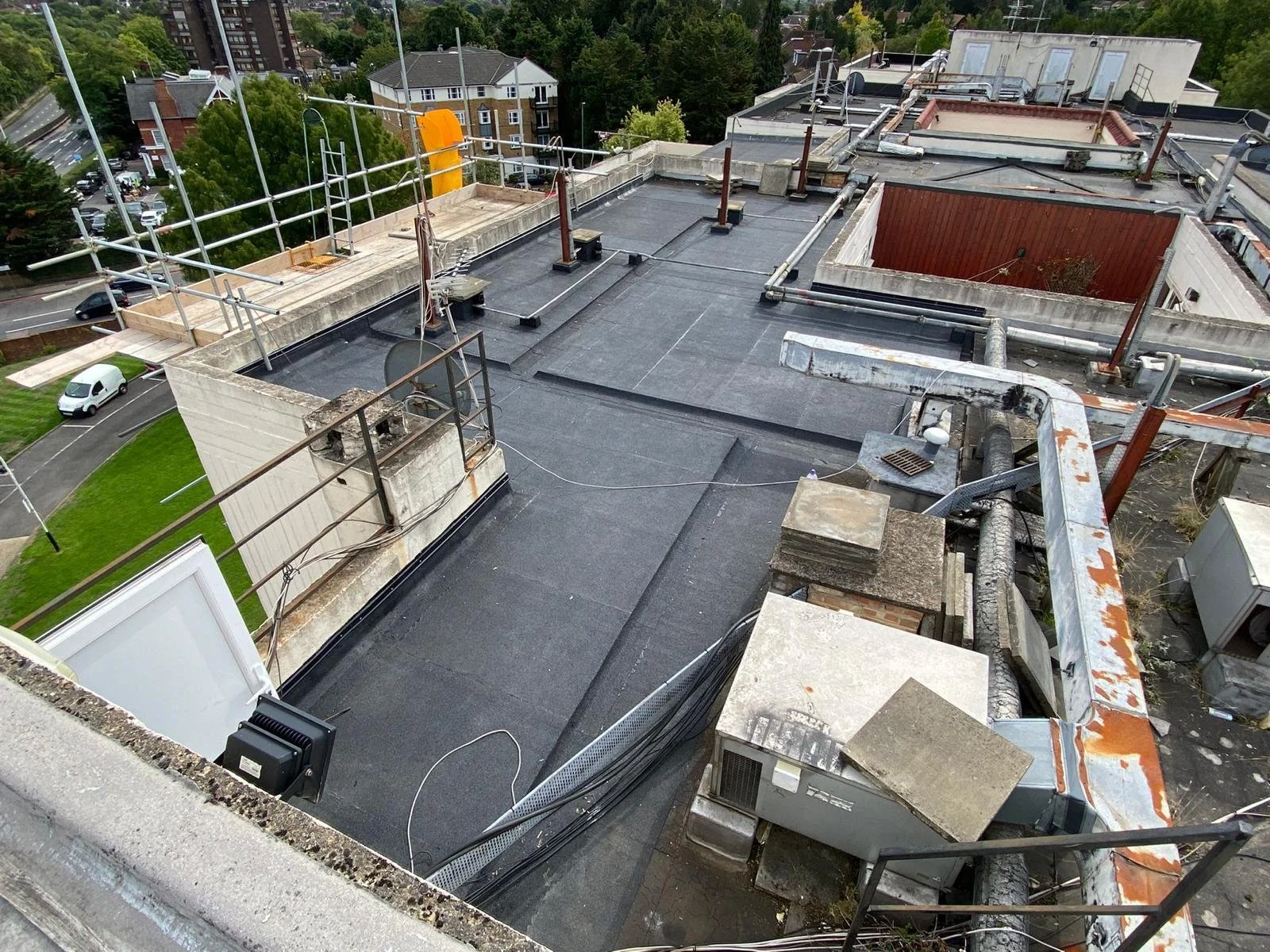 View of a rooftop under construction with scaffolding, various vents, and HVAC equipment, overlooking a neighborhood with trees and buildings.