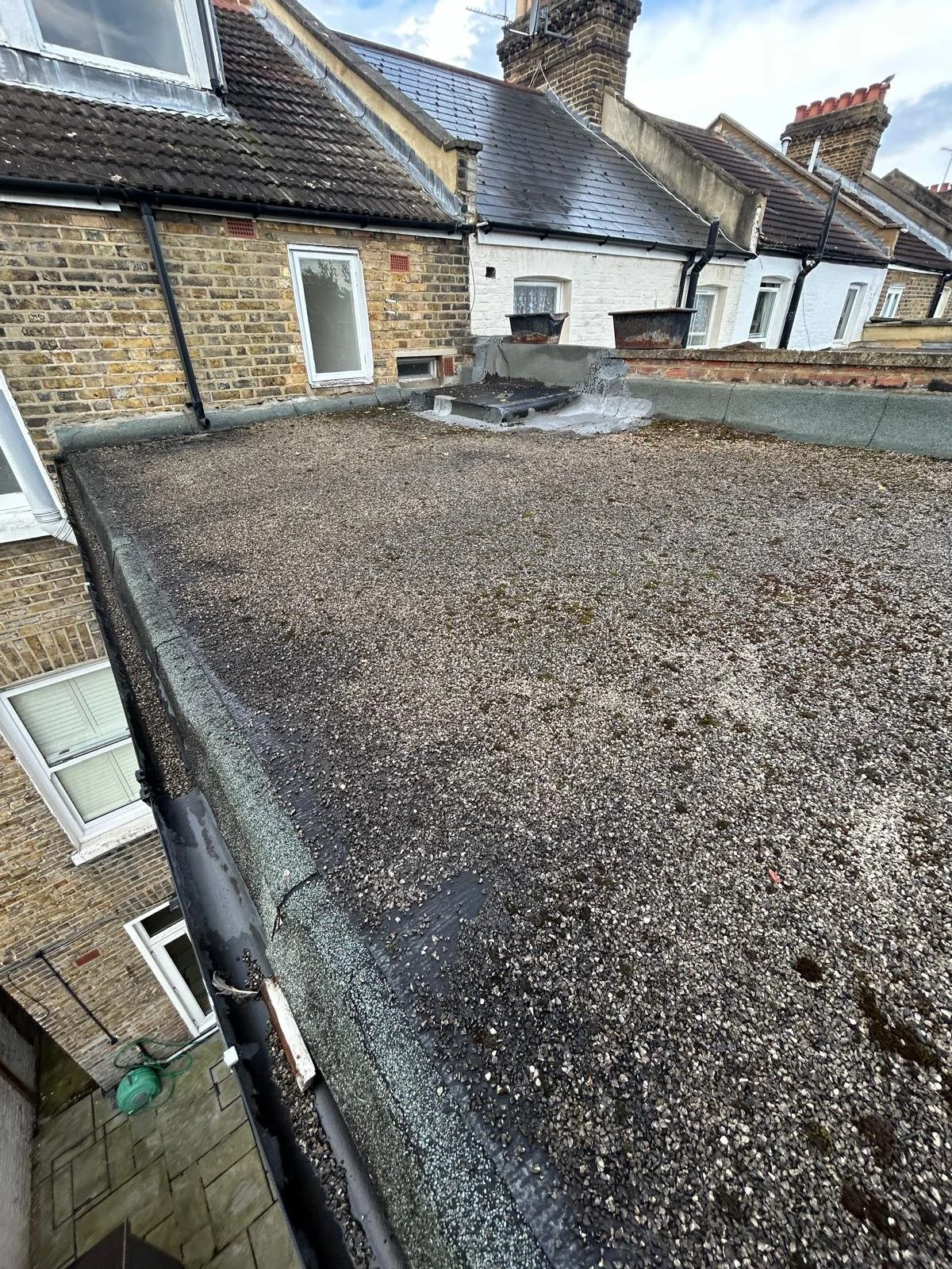 Rooftop with gravel surface, vent pipes, roof flashing, and neighboring brick and white buildings with chimneys.