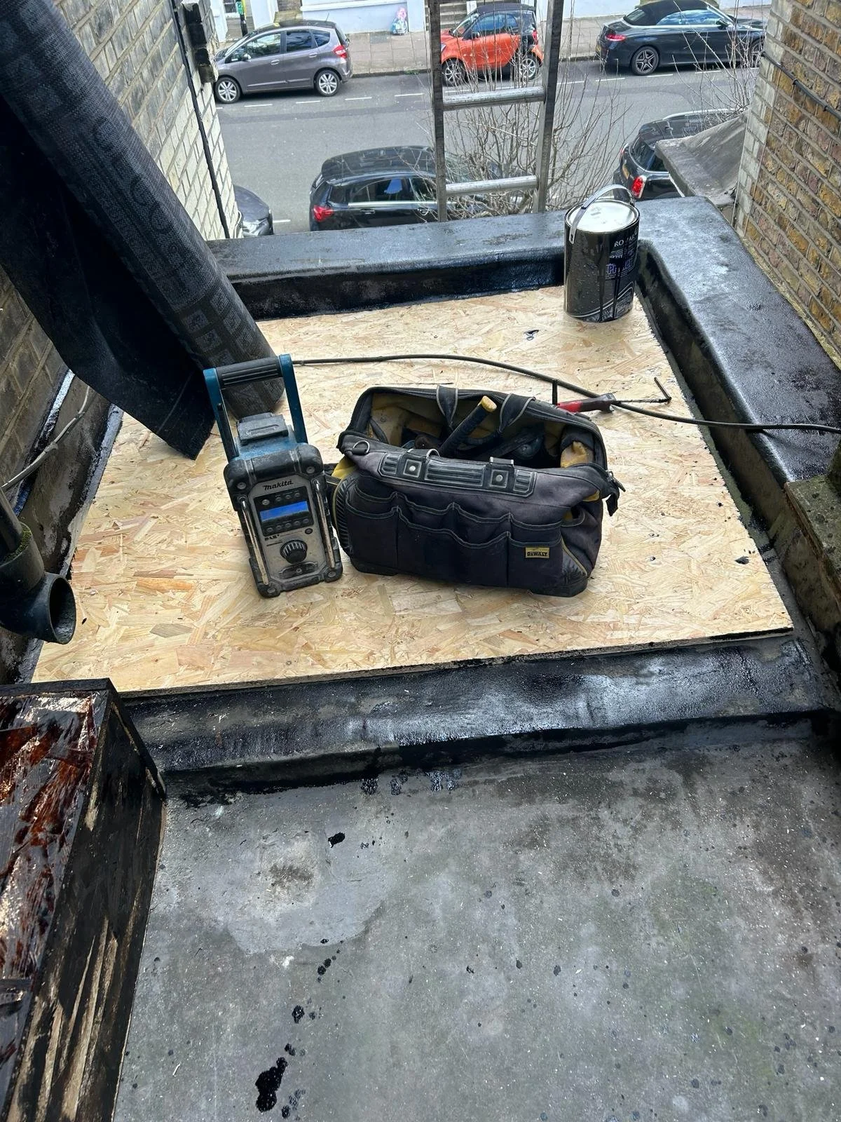 Construction work site with plywood on floor, tools including a tool bag, a measurement device, a paint bucket, and a cord, set on a ledge overlooking a street with parked cars.